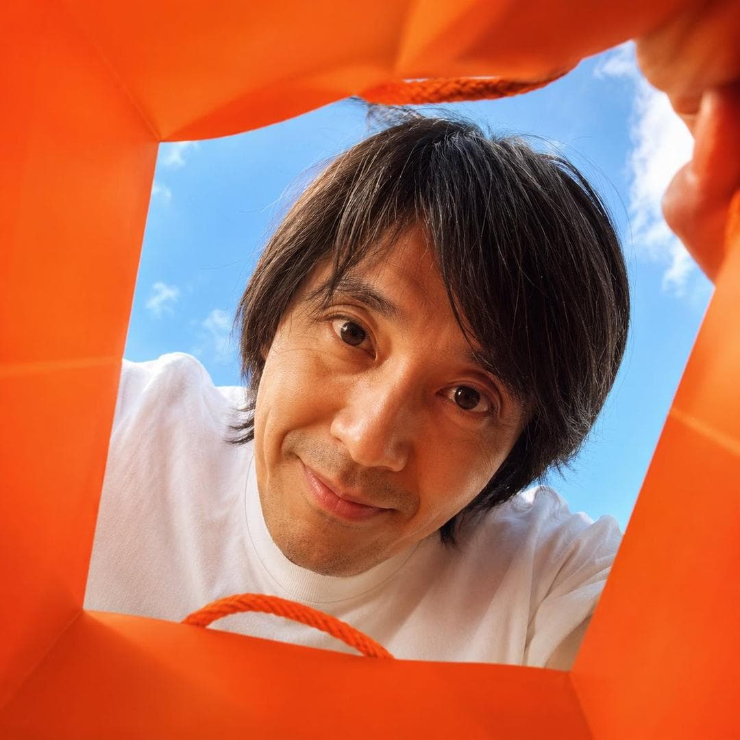 Create a vibrant, adorable, high-end advertising photo shot from inside a shopping bag, looking upward through the wide open top toward the sky. The camera is placed deep inside the shopping bag, using a wide-angle lens at f/1.4 to create a playful close-up perspective with slight but charming wide-angle distortion. This distortion is important and must be clearly preserved, giving the image a fun, immersive, premium-advertising feel. The edges of the shopping bag should naturally frame the image, showing premium paper texture, soft folds, handles, and a clean commercial packaging aesthetic. The shopping bag should be a high-saturation color, with the model randomly choosing a vivid tone such as hot pink, saturated yellow, blood orange, cobalt blue, vivid green, electric purple, deep cyan, or other bold commercial colors. The overall image should feel bright, cheerful, colorful, and visually striking. Main subject: If FACE_REF is uploaded: Strictly preserve the uploaded person’s facial identity with 100% fidelity. Maintain the exact facial structure, eyes, eyelids, nose, lips, jawline, cheekbones, skin texture, age impression, hairstyle tendency, and overall identity. No beautification drift, no face reshaping, no age change, no ethnicity change, and no invented features. If only PERSON_NAME is provided: Create a highly recognizable portrait based on the public image of [周星驰], preserving their iconic facial features, hairstyle, expression tendency, posture, and personal aura as faithfully as possible. The person leans deeply into the shopping…
