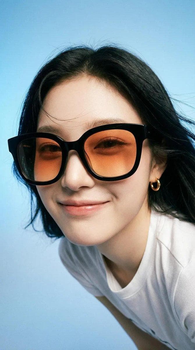 Photorealistic editorial portrait of a smiling woman using the exact same face from the reference image. She wears oversized black sunglasses with orange lenses and small gold earrings. Slightly leaning forward in a close wide-angle perspective, with a playful, mischievous expression and subtle smirk…