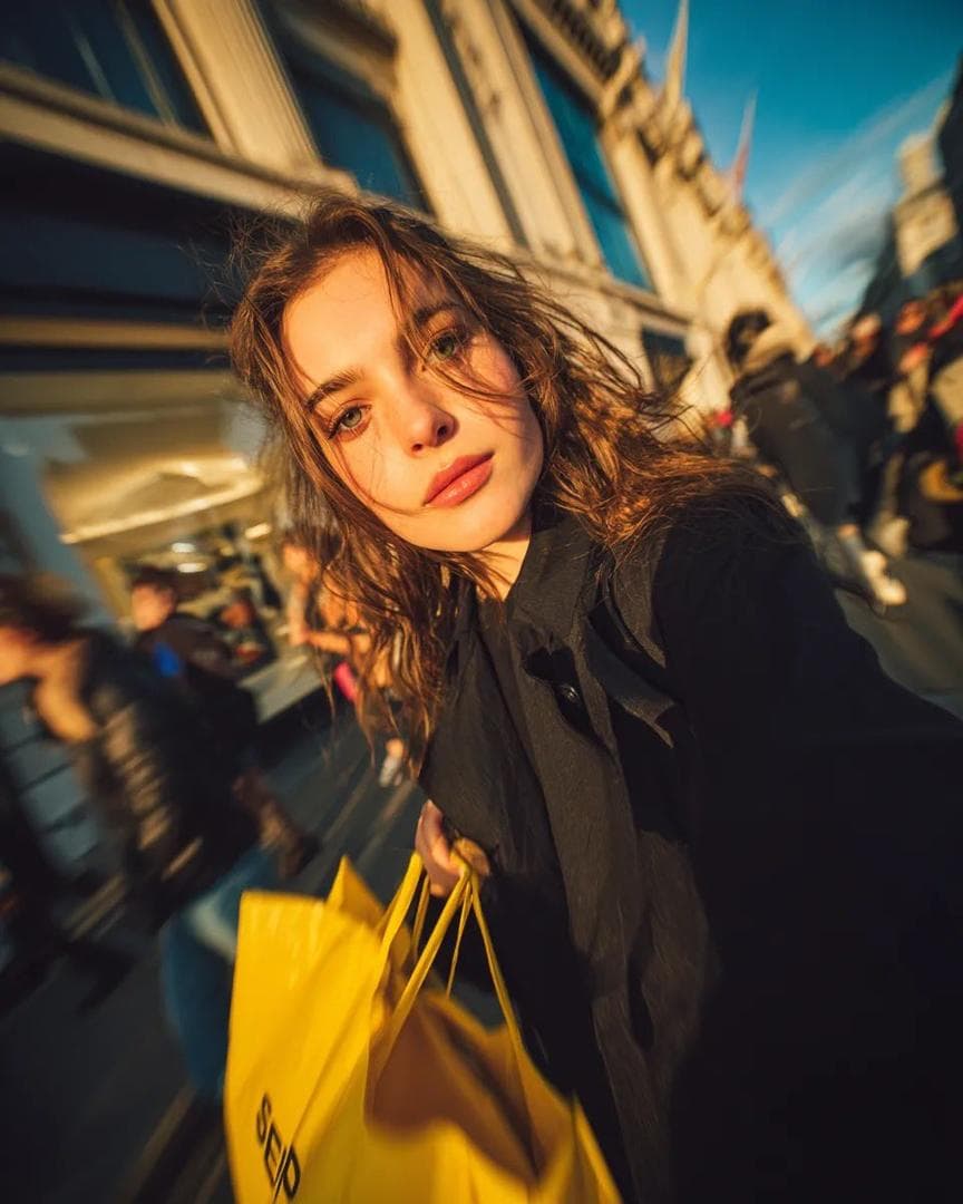 Cinematic street portrait of a young woman walking through a busy shopping street at golden hour, holding bright yellow shopping bags, wind-blown wavy hair, soft natural makeup, intense eye contact with camera, shot from a slightly low angle close-up, dynamic motion blur in the background crowd, sharp focus on face, warm sunlight casting dramatic highlights and shadows…