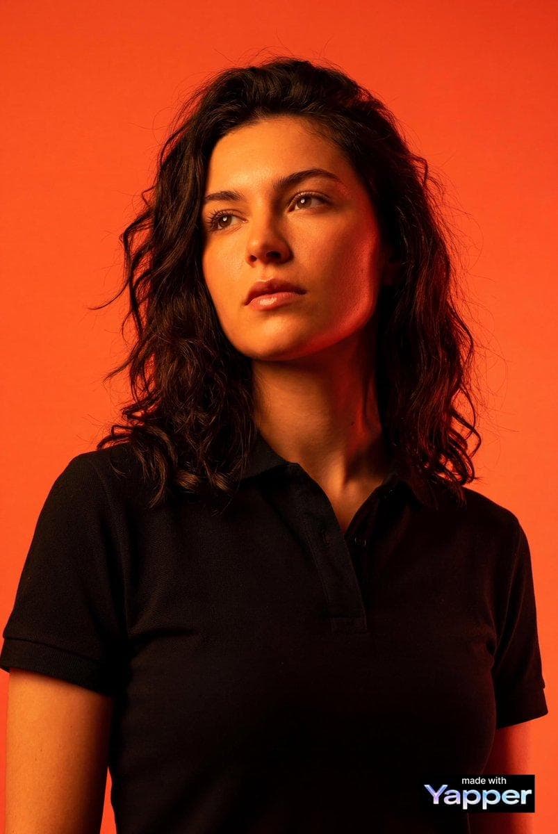 Create a high-impact editorial studio portrait using the person from the attached reference photo as the only identity source. Preserve the subject’s facial structure, skin tone, proportions, hairstyle, age, and overall likeness exactly as in the reference image. Do not modify, enhance, reshape, beautify, or reinterpret any facial features. Absolute identity accuracy is required. The subject is framed in a medium close-up (head and shoulders), photographed from a low-angle perspective looking slightly upward to create a powerful, dominant presence. The subject’s gaze is directed off-camera, upward into space, with a serious, focused, intense expression. Wardrobe: premium black pique polo shirt with visible cotton mesh texture. The fit is sharp and tailored, deep matte black fabric, structured and clean silhouette. Background: solid vibrant orange-red studio backdrop with smooth, seamless color gradients. No patterns or texture — only rich, intense color creating a heated, dramatic atmosphere. Lighting: bold, high-contrast studio lighting dominated by orange and deep red tones. Strong directional key light creates dramatic…
