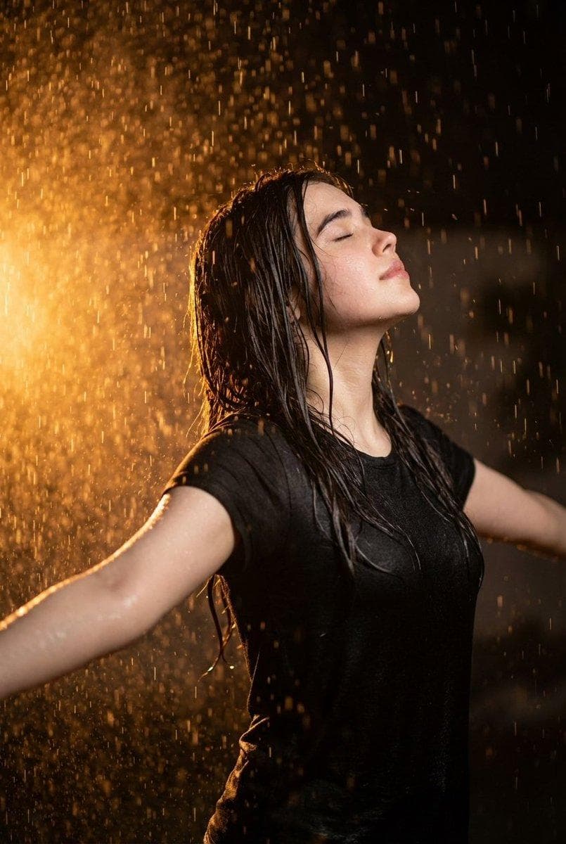 Ultra realistic cinematic portrait of a 32-year-old young woman standing in heavy rain at night, arms open wide and face lifted toward the sky, eyes closed, feeling the rain. Dramatic warm orange backlight illuminating the falling raindrops, creating glowing rain particles around her. Wet black t-shirt tightly fitting on the body, water droplets on her skin. Strong contrast lighting with a dark background, orange and golden light shining from the left side like fire or a spotlight. Emotional expression of freedom and peace…