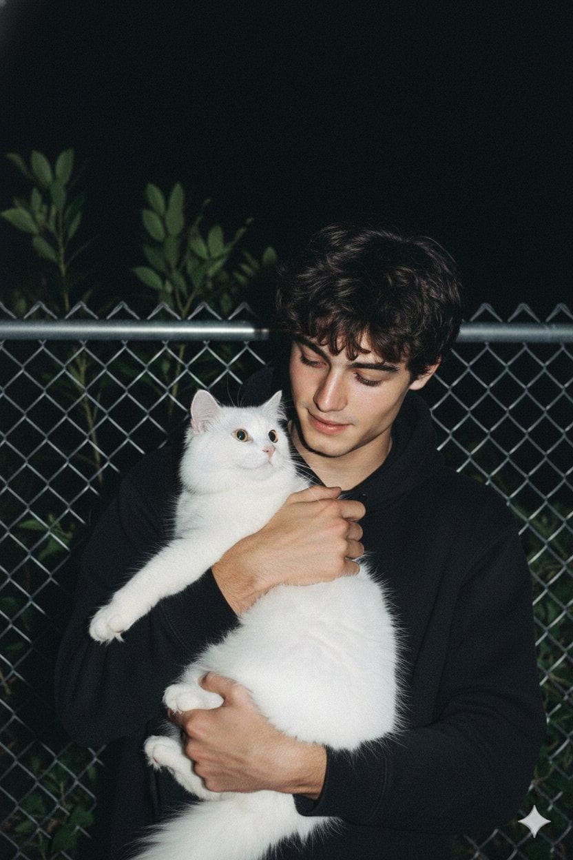 A raw, candid smartphone-style photo taken at night with a soft bright, direct camera flash, use my face on this image, featuring a man with middle-parted dark hair and a soft, gentle expression looking affectionately at a large, fluffy white cat that he is holding in his arms. The man is wearing a plain black hoodie, and the cat is looking back at him, creating a heartwarming moment. The background consists of a metal chain-link fence with some dark green leafy…