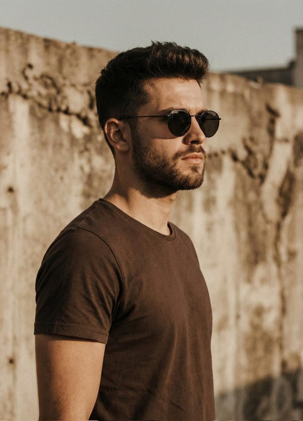 A cinematic portrait of a handsome man with a short styled haircut and trimmed beard, wearing a plain brown t-shirt and round black sunglasses, standing in side profile against a textured concrete wall, warm golden-hour sunlight, shallow depth of field…