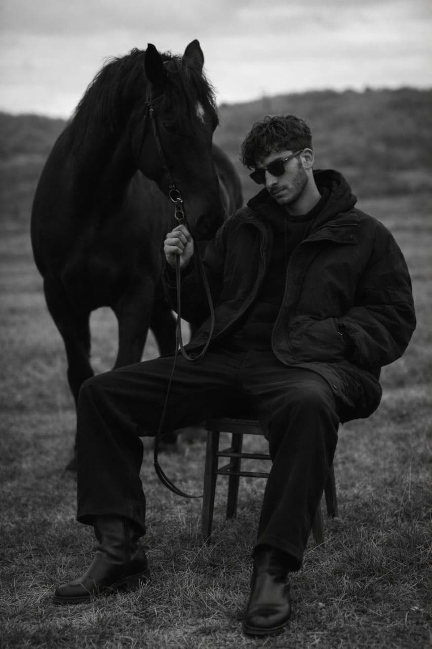 { "model": "Nano Banana Pro", "scene": "Open field landscape under an overcast sky, minimal natural surroundings", "subject": { "type": "Young man", "pose": "Seated on a wooden chair, legs relaxed apart, holding a horse lead calmly", "expression": "Detached, introspective, controlled", "gaze": "Looking downward, partially obscured by sunglasses", "emotion": "Quiet dominance, solitude, inner gravity" }, "appearance": { "hair": { "color": "Dark", "style": "Short, slightly tousled curls" }, "facial_features": { "beard": "Light stubble", "accessories": ["Dark sunglasses"] }, "skin": "Natural texture, matte finish" }, "attire": { "outerwear": "Oversized dark padded jacket", "innerwear": "Dark hoodie", "pants": "Relaxed-fit dark trousers", "footwear": "Black leather boots", "style_note": "Minimalist, masculine, utilitarian fashion" }, "secondary_subject": { "type": "Black horse", "position": "Standing beside the man, head lowered slightly", "interaction": "Held calmly by a loose lead", "symbolism": "Strength, instinct, controlled power" }, "environment": { "location": "Open grassy field", "background": [ "Rolling hills", "Soft distant landscape", "No urban elements" ], "atmosphere": "Quiet, isolated, contemplative" }, "lighting": { "type": "Natural diffused daylight", "conditions": "Overcast sky", "effect": "Soft shadows, low contrast, even tonal range" }, "camera": { "angle": "Eye-level to slightly low-angle", "lens": "50mm cinematic portrait lens", "depth_of_field": "Moderate, subject and horse in focus, background gently blurred", "framing": "Vertical, full-body portrait with symbolic spacing" }…