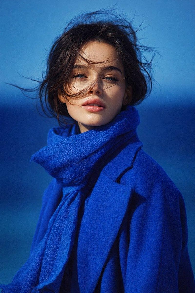 { "image_type": "portrait_photography", "resolution_target": "8K UHD", "aspect_ratio": "vertical", "frame_composition": { "crop": "mid-chest to head", "orientation": "portrait", "subject_position": "centered", "camera_angle": "eye-level", "perspective": "straight-on" }, "subject": { "gender_presentation": "female", "age_appearance": "young adult", "face": { "shape": "oval", "skin_tone": "fair with neutral undertone", "skin_texture": "smooth, natural, visible realism, no heavy retouching", "facial_features": { "eyes": { "color": "light blue-green", "shape": "almond-shaped", "gaze_direction": "directly toward camera", "eyelids": "slightly relaxed", "eyelashes": "natural length, not exaggerated" }, "eyebrows": { "color": "dark brown", "thickness": "medium", "shape": "soft natural arch" }, "nose": { "shape": "straight", "size": "proportionate", "bridge": "smooth" }, "lips": { "color": "soft natural pink", "fullness": "medium to full", "state": "slightly parted" } }, "expression": "neutral, calm, composed" }, "hair": { "color": "dark brown", "length": "short to medium", "style": "loose, slightly wavy", "movement": "wind-blown strands crossing face", "parting": "natural, unstructured" } }, "clothing": { "outerwear": { "type": "coat", "color": "deep royal blue", "material_appearance": "wool or felt-like texture", "fit": "structured but relaxed", "visible_details": "clean seams, minimal design" }, "accessory": { "type": "scarf", "color": "matching royal blue", "material_appearance": "soft, fabric texture", "placement": "wrapped loosely around neck" } }, "color_palette": { "dominant_colors": [ "royal blue", "deep blue", "soft skin tones" ], "contrast": "high contrast between blue clothing and lighter skin", "color_temperature": "cool" }, "lighting": { "type": "natural light", "direction": "frontal with slight side softness", "intensity": "moderate", "shadows": "soft, minimal harsh shadows", "highlights": "subtle…