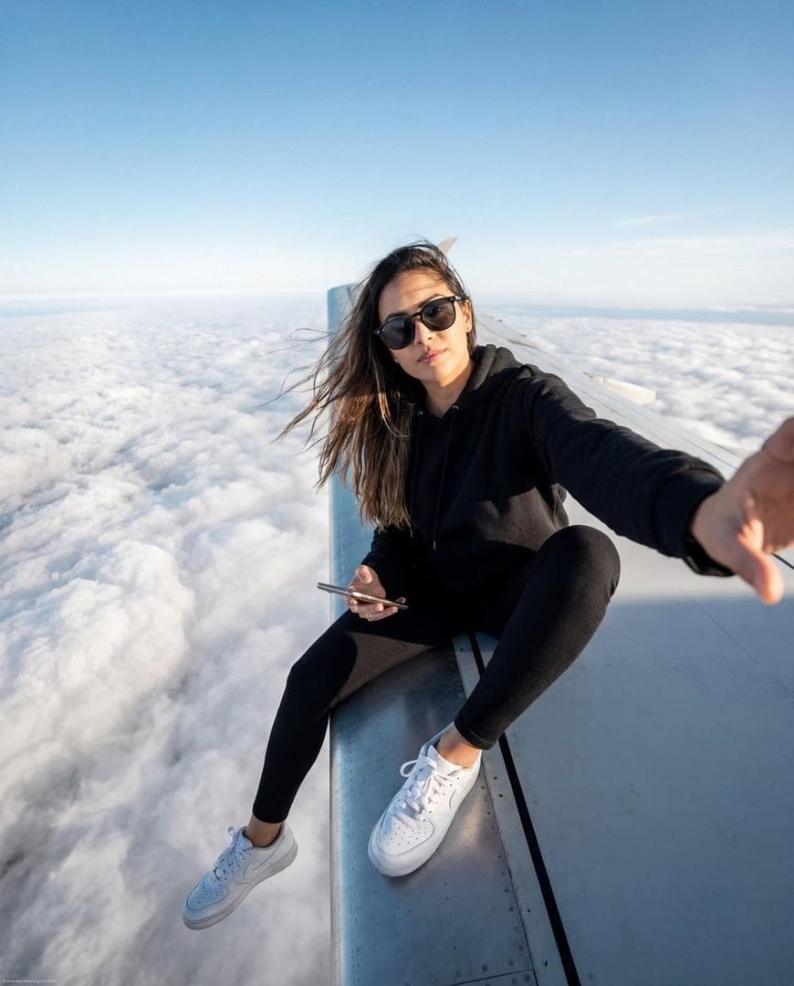 { "image_generation": { "subject": { "description": "stylish young woman sitting on an airplane wing high above the clouds", "pose": "seated casually on the airplane wing, one hand extended forward as if taking a selfie, relaxed but confident posture", "expression": "calm, confident, composed", "gaze": "looking toward the camera with a cool, self-assured expression" }, "appearance": { "hair": { "style": "long, natural, slightly flowing in the wind", "look": "casual and effortless" }, "accessories": [ "dark sunglasses", "minimal jewelry" ], "vibe": "adventurous, bold, modern" }, "outfit": { "style": "sporty casual", "details": "black hoodie, black fitted pants, clean white sneakers" }, "environment": { "setting": "high above the clouds on an airplane wing", "sky": "clear blue sky with soft clouds below", "atmosphere": "open, airy, dramatic aerial view" }, "lighting": { "type": "natural sunlight", "style": "bright and cinematic", "direction": "side lighting from the sun", "quality": "crisp highlights with soft shadows" }, "camera": { "angle": "wide selfie-style perspective from a slightly elevated angle", "lens": "ultra wide-angle"…