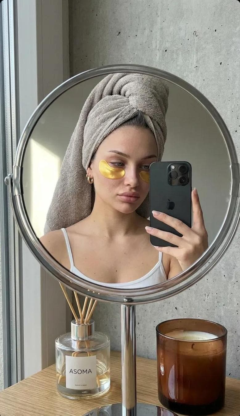 A highly realistic mirror selfie of a young woman taken through the reflection of a round tabletop mirror with a chrome metal frame and stand. The composition feels intimate and natural, like a quiet personal care moment. She is wearing a thin white spaghetti-strap tank top. Her head is wrapped in a soft grey-beige terry towel styled as a turban, fully covering her hair. Under her left eye, a single pair of gold hydrogel eyepatches is applied — semi-circular, slightly reflective with a subtle metallic sheen. Her face appears makeup-free or with very minimal natural makeup. Skin looks fresh, hydrated, and real, with visible natural texture, soft pores, slight tonal variations, and gentle under-eye shadows. Lips have a neutral tone with a light natural gloss. She wears small gold hoop earrings. She holds a black iPhone with a triple-camera module (similar to iPhone 11 Pro or newer) in her right hand, angled toward the mirror. Her expression is calm and relaxed, slightly tired but peaceful, with her gaze directed slightly downward at the phone screen. Environment & background: Behind her is a grey concrete-textured wall. On the left side of the frame, a window allows soft natural daylight to enter, creating gentle, diffused illumination with a slightly cool tone. On the table in front of the mirror: On the left: a clear glass diffuser bottle with wooden sticks, labeled “ASOMA”. On the right: a brown…