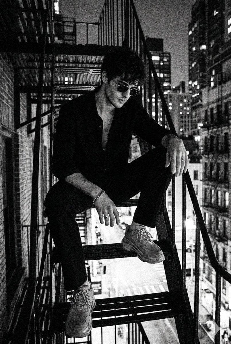 Create a portrait without changing the facial features. A black and white artistic photograph of a man in a noir style. The man is sitting on a metal staircase or balcony with a cityscape in the background. He is wearing a dark shirt with an open collar, dark trousers, chunky sneakers, and sunglasses. Accessories are visible on his hands, including a bracelet. His pose is relaxed, with his gaze directed downward. The lighting is high contrast with deep shadows. The atmosphere…