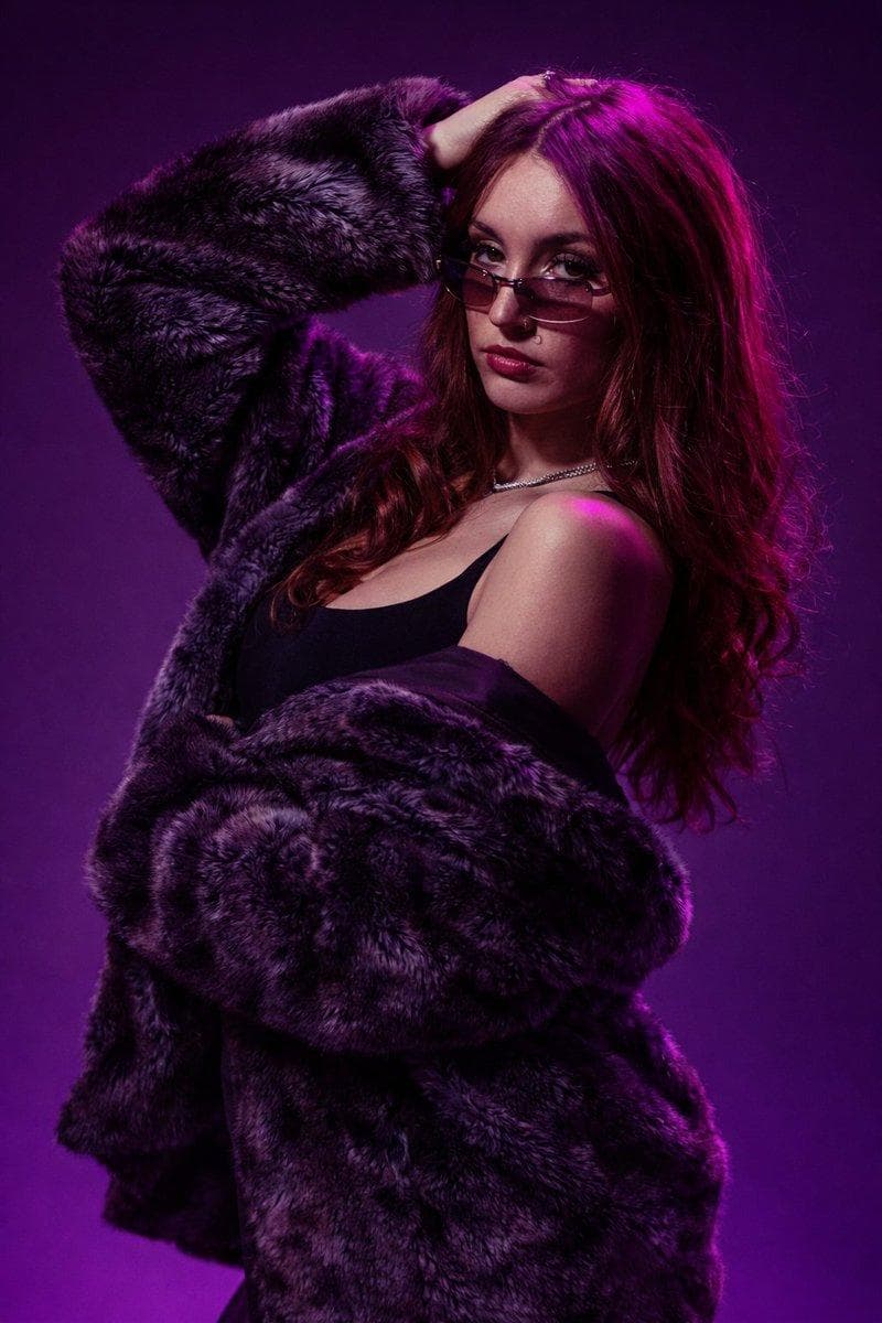 { "image_type": "studio fashion portrait photography", "resolution_target": "8K ultra high definition", "aspect_ratio": "portrait (vertical)", "composition": { "framing": "medium portrait", "crop": "head to mid-torso", "subject_position": "slightly right of center", "camera_angle": "eye-level", "pose": { "body_orientation": "three-quarter turn to the left", "head_orientation": "turned toward camera", "arm_position": "one arm raised with hand placed behind or on top of head, other arm lowered and relaxed" } }, "subject": { "gender_presentation": "female", "age_range": "young adult", "skin_tone": "light to medium", "facial_expression": { "expression": "neutral to mildly serious", "gaze_direction": "directly at camera", "emotion": "confident, composed" }, "face_details": { "face_shape": "oval", "complexion": "smooth, even", "makeup": { "eyes": "defined eyeliner and mascara", "lips": "deep muted red or berry tone lipstick", "overall": "moderate, polished makeup" }, "piercings": [ "small nose ring on left nostril" ] }, "eyes": { "visibility": "partially visible through tinted glasses", "eyewear": { "type": "narrow rectangular sunglasses", "lens_color": "dark tinted", "frame": "thin metal frame", "position": "worn low on the bridge of the nose" } }, "hair": { "color": "medium brown with strong magenta/purple lighting highlights", "length": "long", "style": "loose, softly wavy", "parting": "center part" } }, "clothing_and_accessories": { "outerwear": { "type": "faux fur coat", "color": "dark gray to black", "texture": "thick, plush, high-volume fur", "fit": "oversized", "wearing_style": "draped loosely off the shoulders" }, "inner_clothing": { "type": "sleeveless top", "color": "black", "neckline": "straight or slightly scooped" }, "jewelry": { "necklace": { "type": "thin chain necklace", "material": "metallic", "visibility": "clearly visible at collarbone" } } }, "lighting": { "style": "studio…