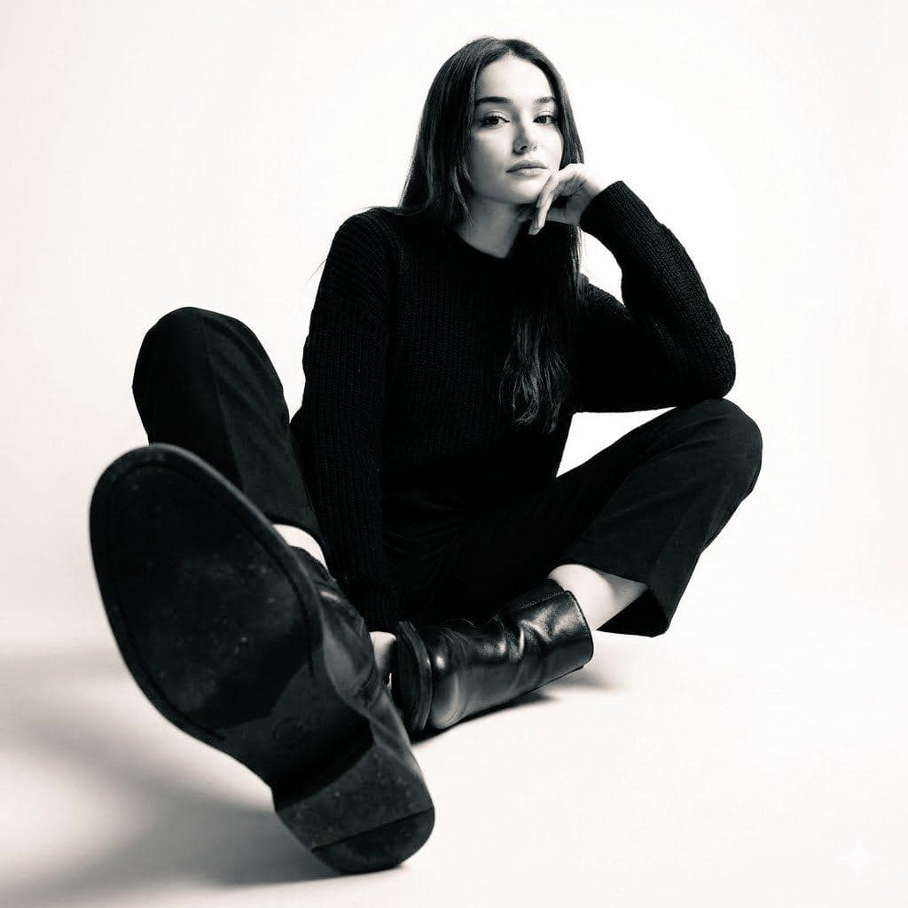 High-contrast black-and-white studio photo of a woman seated on the floor wearing a black sweater, black trousers, and black leather ankle boots. Legs are bent and open toward the camera, with one boot sole prominently facing the lens. Upper body slightly reclined…