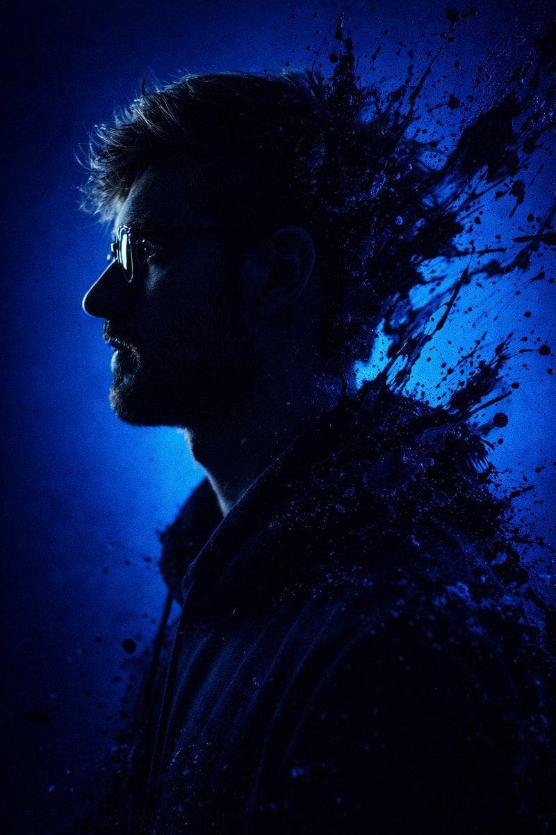 Convert this image into a dramatic, stylized portrait (use the same face as the uploaded photo), 9:16 aspect ratio, featuring a dark, almost silhouetted figure in profile looking to the left. The background is a deep electric blue. Intense black and sapphire paint splashes burst outward…
