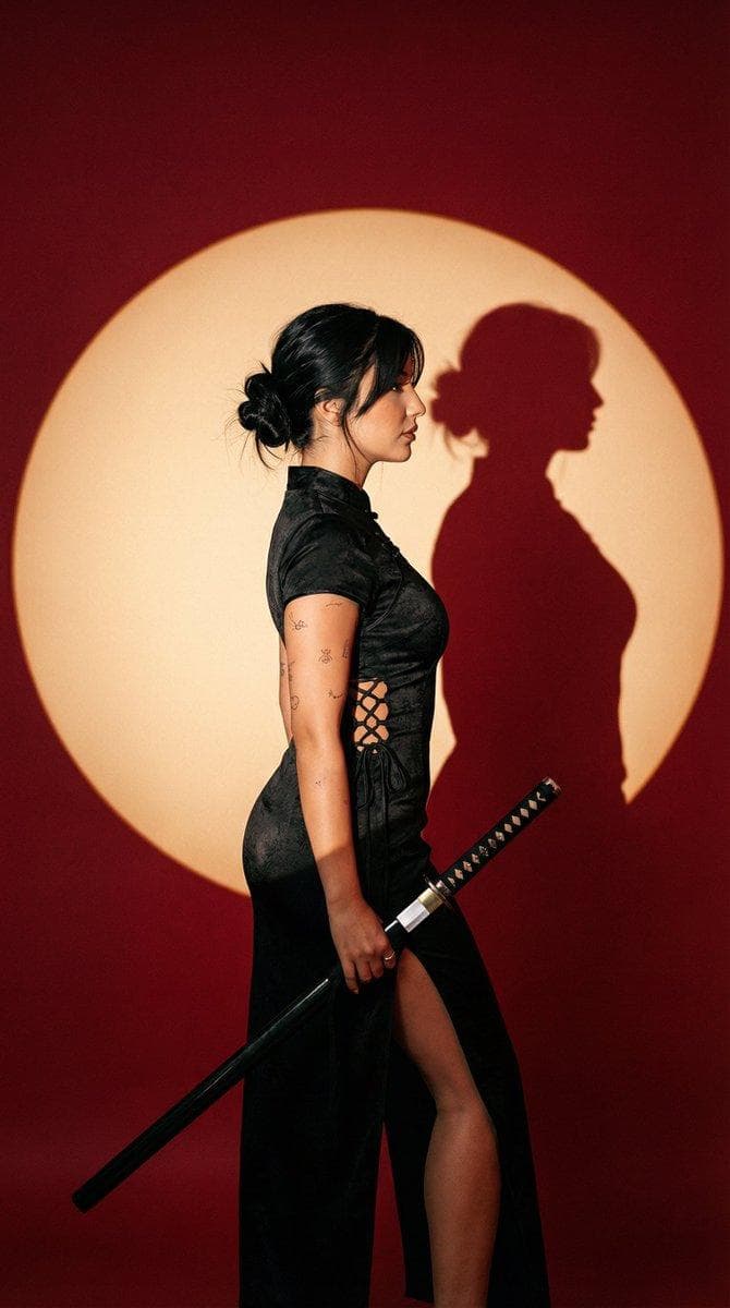 { "generation_request": { "meta_data": { "task_type": "photoreal_editorial_spotlight_disk_fashion_portrait", "language": "en", "priority": "highest", "version": "v1.0_RED_BACKDROP_CIRCLE_SPOTLIGHT_KATANA_EDITORIAL" }, "output": { "aspect_ratio": "4:5", "resolution": "high", "num_images": 4 }, "scene": { "concept": "minimal graphic studio portrait with a circular spotlight disk and crisp shadow silhouette", "environment": "deep red seamless background, no props, no clutter", "composition": "3/4 body or full-body, side profile; subject placed slightly right of center; large circular spotlight disk behind subject" }, "subject": { "person": "adult woman", "wardrobe": { "dress": "fitted black cheongsam-inspired dress, high slit, lace-up side detailing, matte fabric with subtle texture" }, "hair_makeup": { "hair": "messy low bun, a few loose strands framing face", "makeup": "soft glam, defined eyeliner, natural lips, realistic skin texture" }, "details": { "tattoos": "subtle small tattoos on upper arm, realistic ink" }, "pose": "side profile stance, shoulders relaxed, one hand resting near hip, holding a katana angled downward in a safe, non-aggressive pose", "expression": "calm, composed, editorial" }, "props": { "katana": { "style": "realistic katana with correct proportions, simple handle wrap, subtle metal sheen", "handling_notes": "safe grip, blade angled down, no threatening gesture" } }, "lighting": { "style": "single hard key light + spotlight gobo/disk", "notes": "create a large circular warm spotlight disk (cream/peach) on the red background; crisp hard-edged shadow silhouette of subject visible…