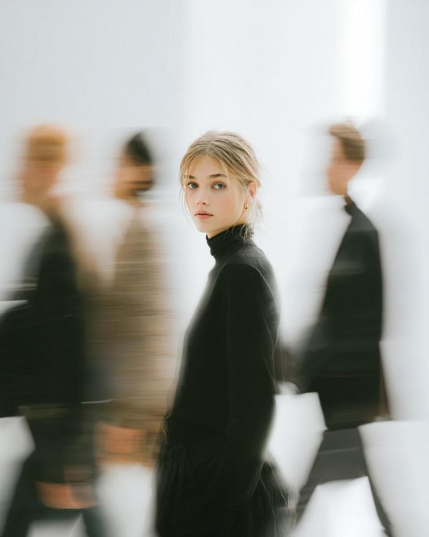A high fashion magazine pictorial of an Alpine-looking woman in her early 20s who stands still, looking at the camera, turning her head in the center, surrounded by a blurry silhouette of people passing by. She has a calm, stoic look on her face and is wearing a black minimalist outfit. The background is clean, bright and slightly abstract, and emphasizes…