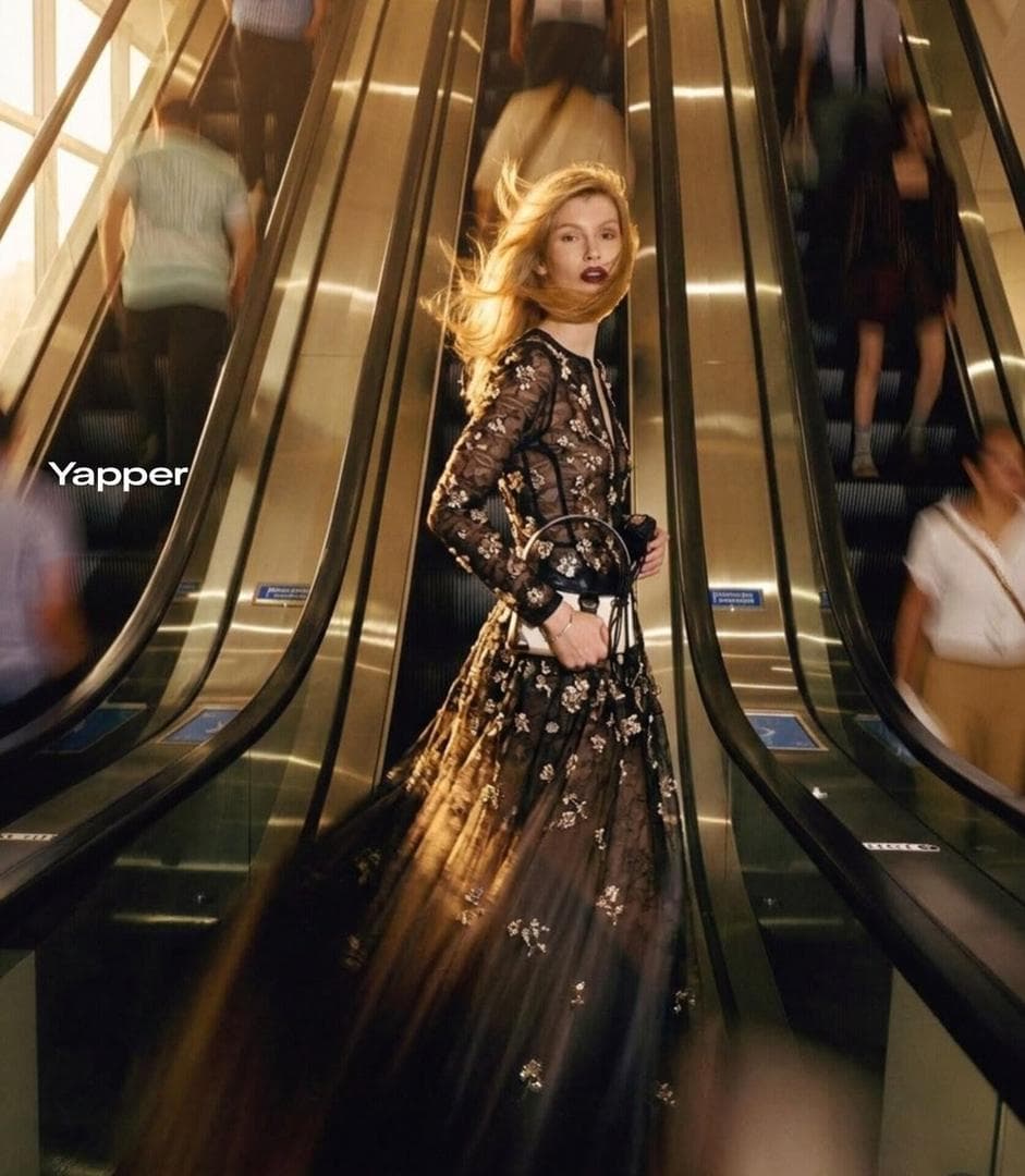 Ultra-realistic cinematic fashion photograph of a stylish blonde woman standing confidently between two moving escalators inside a luxury shopping mall, wearing a sheer black lace long-sleeve top and flowing skirt, holding a small white designer handbag, her hair dramatically blowing sideways as if caught in motion, blurred crowd moving up and down the escalators creating dynamic motion streaks around her, she remains sharp and perfectly focused in the center, dramatic fashion editorial…