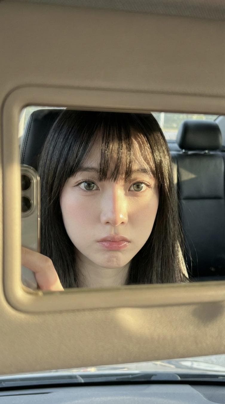{ "meta": { "aspect_ratio": "9:16", "quality": "ultra_photorealistic", "resolution": "8K", "camera": "iPhone front camera", "lens": "24mm", "style": "raw iphone realism, pinterest girl aesthetic, natural skin texture, zero plastic look" }, "composition": { "concept": "frame within a frame", "framing": "extreme close-up face reflection inside car sun visor vanity mirror", "mirror_frame": { "material": "thick beige / cream plastic", "visibility": "dominant around edges of photo, slightly worn texture" }, "phone_visibility": { "type": "iPhone", "detail": "partial phone edge visible on left side of mirror, controlling the shot", "camera_bump": "subtle, realistic" } }, "scene": { "location": "inside parked car", "environment": [ "dark car interior", "black leather seats", "headrest visible in reflection" ], "time": "golden hour", "atmosphere": "quiet, intimate, candid daily life moment" }, "lighting": { "type": "direct golden hour sunlight", "direction": "sharp side sunlight from afternoon sun", "color_temperature": "warm golden", "contrast": "high", "effects": [ "strong highlights on skin", "abstract hair shadow patterns across forehead and cheek", "natural light bloom on lips and eyes" ] }, "subject": { "gender": "female", "age": "early 20s (clearly adult)", "ethnicity": "East Asian / mixed pale tone", "face_structure": { "shape": "soft oval", "cheeks": "full, youthful, slightly flushed", "jawline": "delicate and smooth", "nose": "small, straight, soft rounded tip" }, "eyes": { "size": "large", "shape": "round-almond hybrid", "color": "grey-green / light hazel", "vibe": "doll-like, calm, distant", "lashes": "long, natural, softly separated", "under_eye": "subtle natural shadow for soft tired-cute look", "gaze": "looking directly into mirror/lens" }, "lips": { "size": "full", "shape": "soft cupid’s bow", "color": "natural pink", "finish": "slightly glossy", "state": "relaxed, faint pout" }, "skin": { "tone": "very pale porcelain", "undertone": "cool pink", "texture": "real skin with visible pores", "finish": "natural glow…