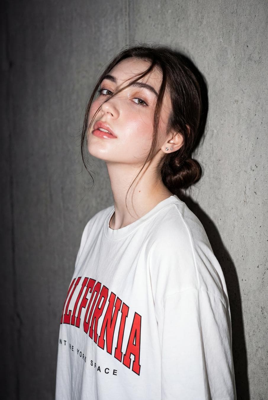 A high-resolution, close-up editorial-style portrait of a young woman standing against a textured gray concrete wall, captured with direct on-camera flash that creates a raw, moody, high-contrast aesthetic. She is positioned slightly angled to the side with her head tilted back and chin lifted, giving an effortlessly cool, candid expression. Her face has a fair, smooth complexion with a natural sheen from the flash, soft rosy blush across the cheeks, minimal eye makeup, lightly defined brows, and glossy natural pink lips that are slightly parted, conveying a relaxed yet confident mood. Wispy strands of her dark brown hair fall loosely across her face, partially covering one eye, while the rest of her hair is pulled back into a low, casual style, enhancing the spontaneous, fashion-editorial feel. Her eyes look slightly upward and past the camera, adding to the detached, artistic vibe. She is wearing a loose-fitting white oversized T-shirt with bold red typography reading “CALIFORNIA” across the chest, along with smaller black text underneath, giving a casual streetwear aesthetic. Small, subtle stud earrings are visible, adding minimal detail without distracting from the overall look. The lighting is harsh and frontal, producing sharp shadows behind her on the wall and emphasizing skin texture, hair strands…