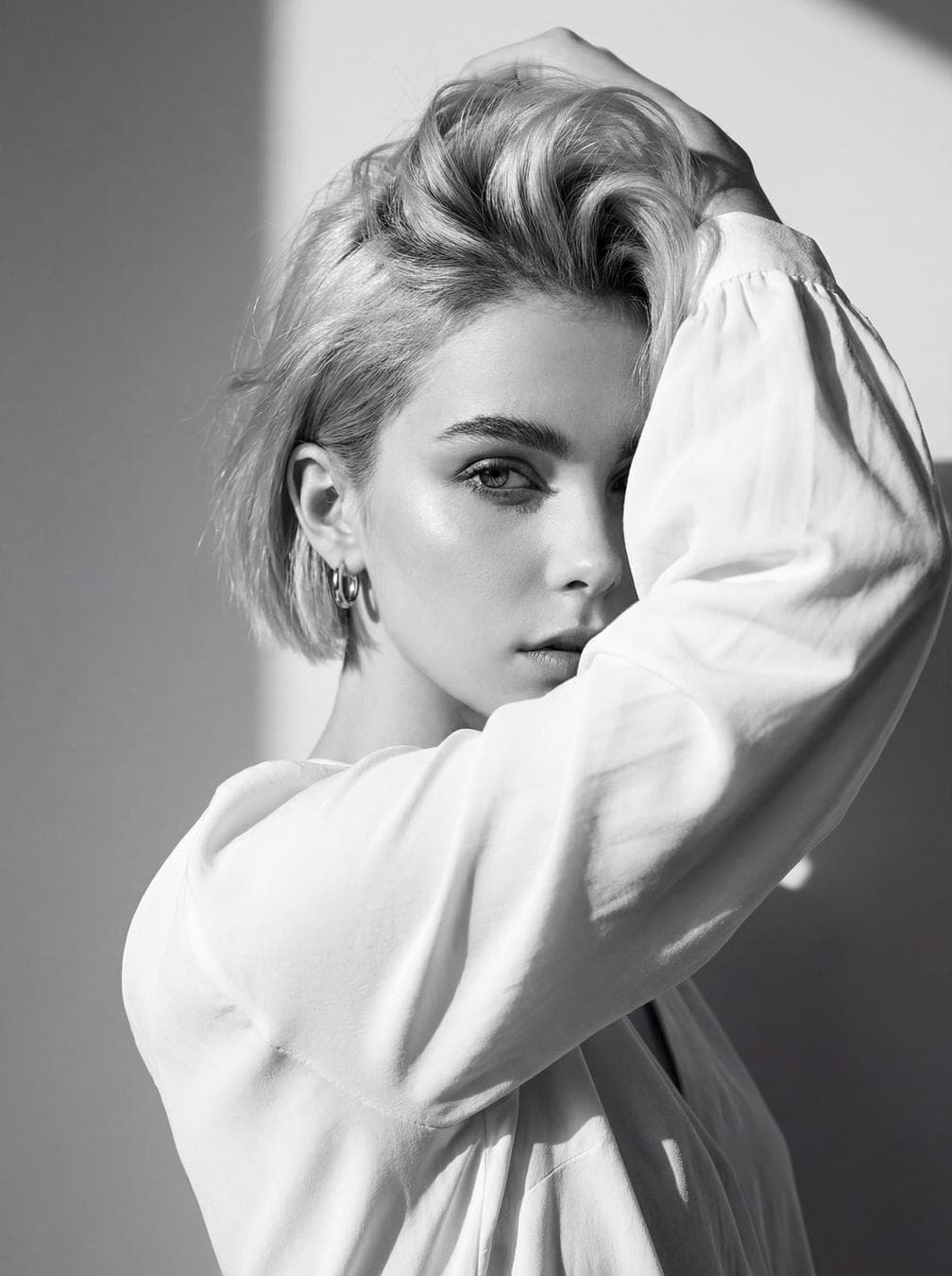 Use the attached image of the model as facial reference and physical features. Black and white high-fashion editorial portrait of uploaded person, wearing a loose white blouse. She poses with one arm raised, partially covering her face while holding her hair up, creating a dramatic and artistic composition. Her gaze is intense and captivating, directed toward the camera, exuding a strong, enigmatic aura. Background: minimalist plain wall with sharp natural daylight casting defined shadows, adding depth and contrast. Lighting: harsh natural sunlight, high-contrast setup highlighting facial contours, sharp cheekbones, and texture of the hair and fabric. Pose: bold and expressive, arm lifted framing the…