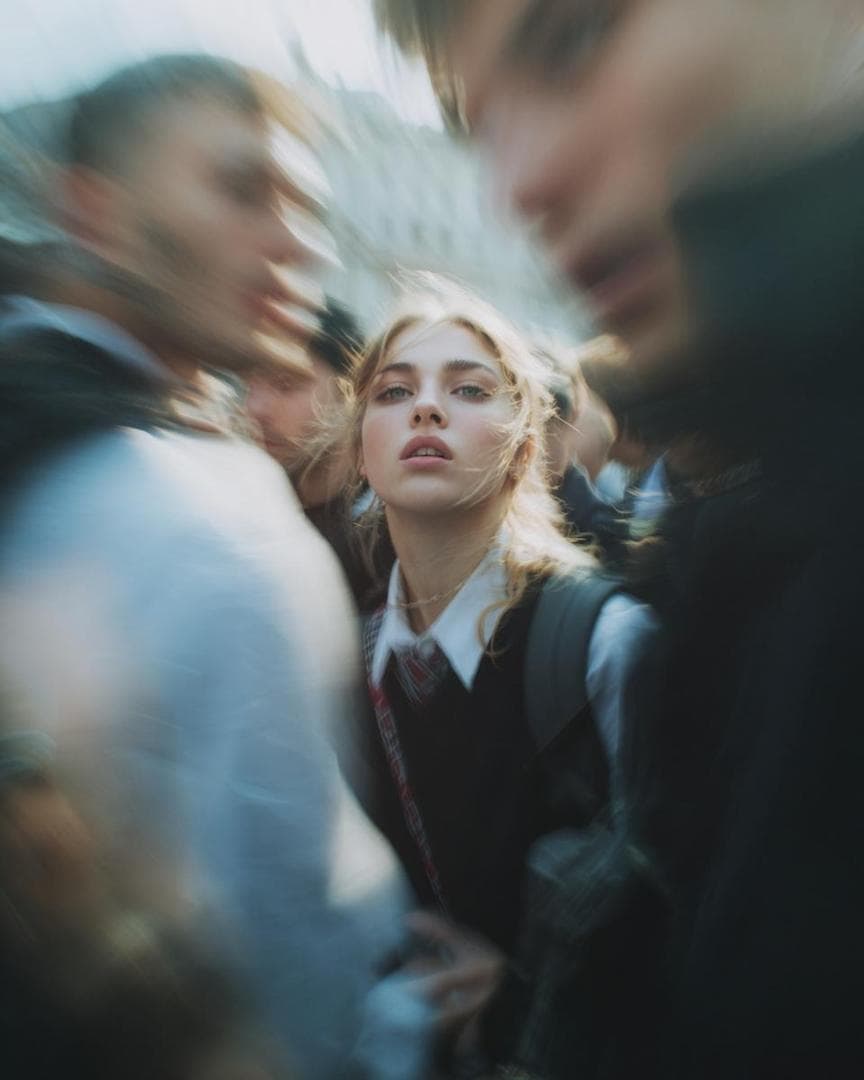 Paparazzi-style extreme close-up photo of a woman with striking facial features, caught off-guard while turning toward the camera. Face and shoulders only, shot from a low angle. Strong harsh on-camera flash, grainy high-ISO, raw candid street-photography feel. Background shows a crowded scene with motion blur (Paris Fashion Week atmosphere). Intense, spontaneous energy, imperfect and real. She is wearing a school uniform. Ultra-realistic, cinematic realism, high detail skin…