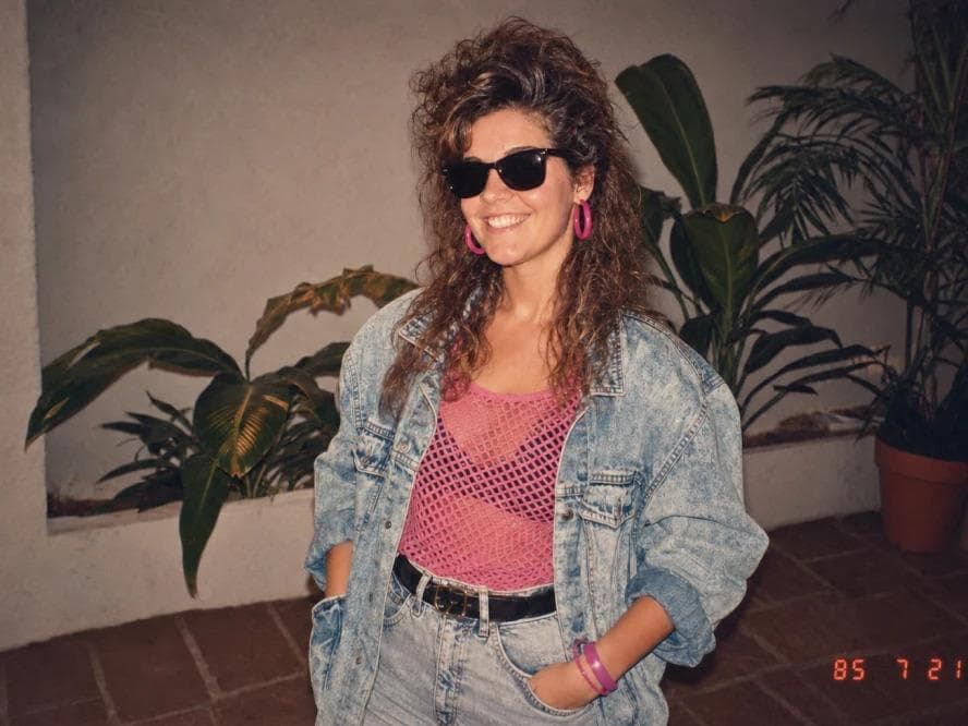 Using my uploaded photo, show me what I would have looked like around 1985. Preserve my identity, facial features, skin tone, age, and recognizable appearance. Reimagine my hair, clothing, accessories, and surroundings with bold, unmistakably mid-1980s styling—expressive silhouettes, statement accessories, layered details, distinctive colors, and textures. Make it feel like a genuine 1985…