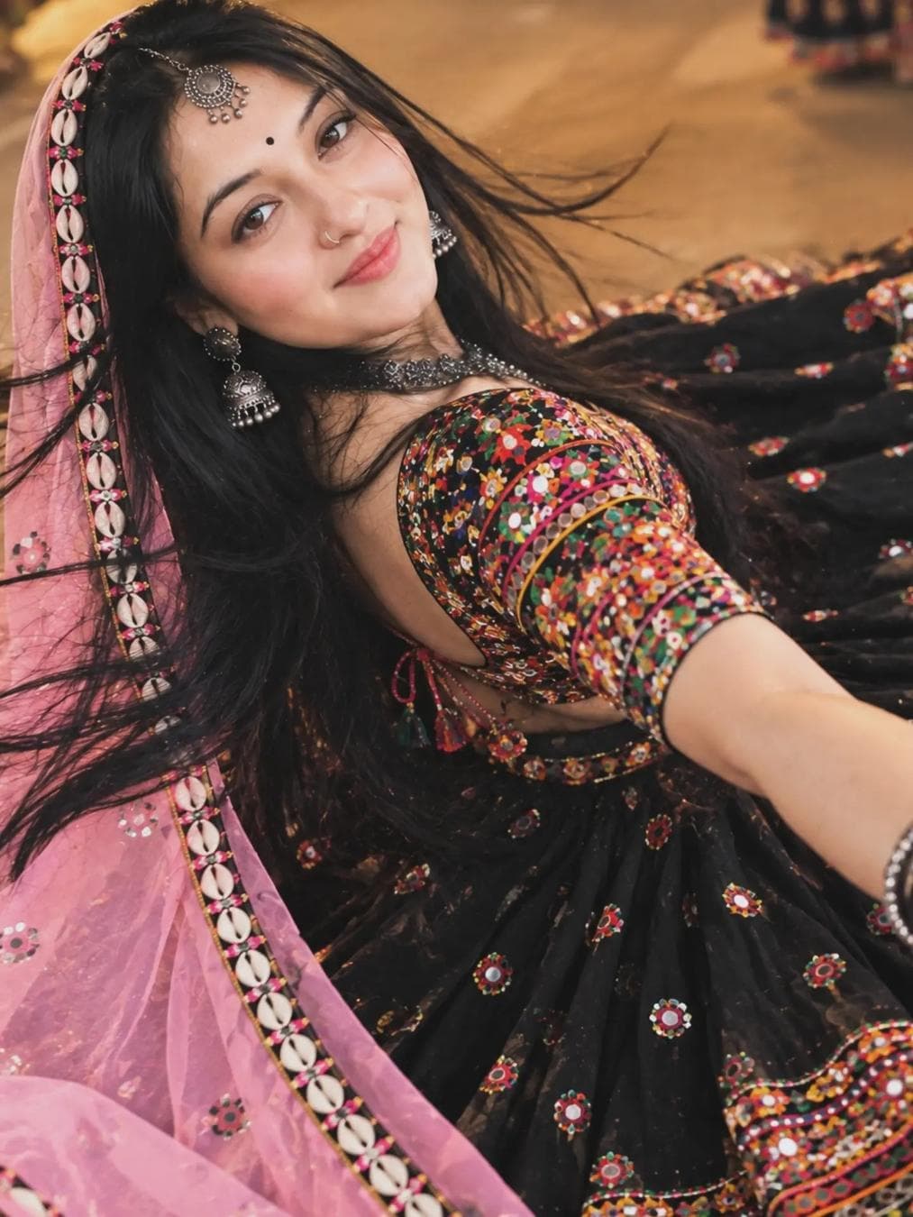 Keep the face exactly the same as the reference photo, with the same facial features and natural appearance. Create a graceful Gujarati garba look with a black, heavily flared lehenga/ghagra featuring beautiful traditional embroidery and mirror work. Pair it with a vibrant multicolored embroidered blouse that creates a striking contrast with the black lehenga. Add a light pastel-pink sheer dupatta draped over the head, starting slightly above the forehead, with an elegant border completely decorated with cowrie shells. Keep the hair long, open, and naturally flowing in the air as she spins during garba. Add a perfect small bindi, a traditional oxidized-silver maang tikka, large oxidized jhumkas, a full oxidized-silver necklace set, and matching bangles. Pose her as if she is gracefully twirling during garba, with the…