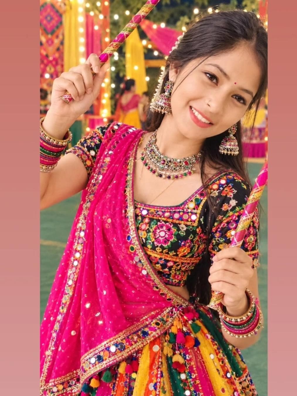 Use my uploaded photo as the exact face reference. Keep the persons original face and identity completely unchanged and recognizable. Do not recreate, replace, reshape, or alter the face, eyes, eyebrows, nose, lips, skin tone, or natural facial features. Create a beautiful full-body Navratri Dandiya/Garba look. Make the head and face straight and naturally aligned, facing the camera. Dress her in a colorful traditional Gujarati mirror-work chaniya choli/lehenga, with a bright pink dupatta, colorful embroidery and traditional detailing. Add beautiful jhumka earrings, a traditional necklace, bangles, small bindi and subtle festive makeup. Give her two decorated Dandiya sticks in her hands in a natural dancing pose. Set the background at a beautiful Navratri Garba venue with colorful pink and yellow drapes, warm fairy lights, decorative lanterns, traditional Gujarati decorations and softly…
