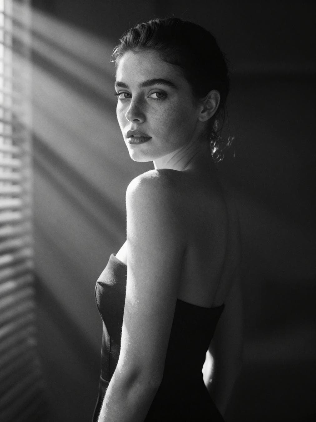 A hyperrealistic 8k black and white cinematic mid shot portrait of a beautiful woman with natural freckles and soft full lips. She is framed from the waist up, standing slightly angled to the camera with one shoulder forward, her head turned back toward the lens in a calm yet evoxative pose. One arm rest slightly against her body while the other drapes loosely, creating a sense of natural elegance. soft beams of sunlight steam through blinds, diffused and hazy, falling across her skin and outfit in blurred patterns of light and shadow. the ray spread like underwater reflection, but glow with a dreamlike softness, sculpting her features gently instead of sharply. Her skin is captured in lifelike texture -- visible pores, delicate freckles, and subtle imperfection -- with the interplay of light emphasizing the natural contours of her face and collarbones. The lips are highlighted with a…