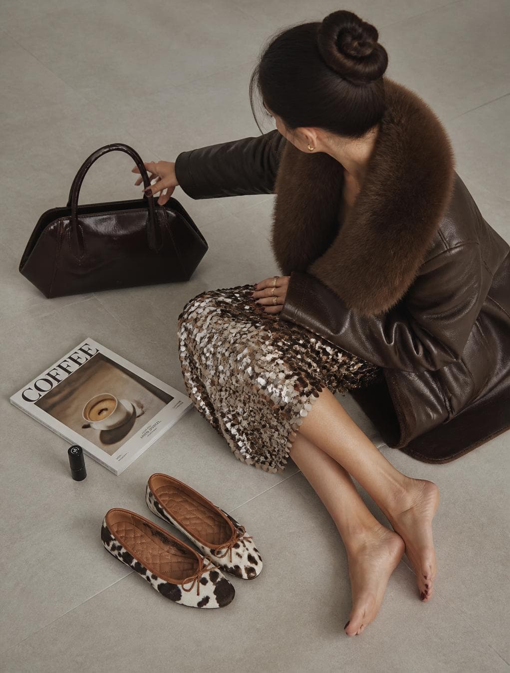 A high-angle shot captures a woman, appearing to be a young adult, with fair skin, seated on a light gray tile floor. Her dark hair is neatly styled in a bun. She wears a brown leather coat with a substantial fur collar, adding an element of luxury. The woman is facing a dark brown, glossy handbag with angular handles. She is barefoot and dressed in a sequined skirt adorned with silver discs, creating a textured contrast with the matte coat. In front of her are a pair of flat shoes with a distinctive black and white animal print and brown details, placed near a coffee-themed magazine labeled "COFFEE" and a small black cylinder, likely a cosmetic product. The overall color palette is neutral, dominated by earthy browns…