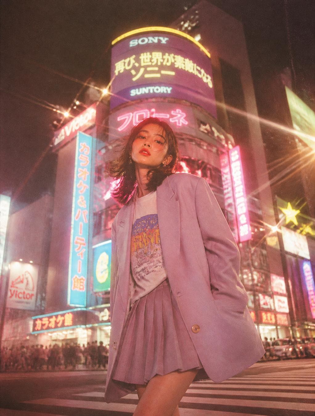 A dreamy cinematic photo of a young Japanese woman in her early 20s standing at a Tokyo pedestrian crossing at night, wearing an oversized pastel blazer over a graphic tee and pleated mini skirt, hair slightly wind-blown. Glowing neon signs in soft pink, cyan, and lavender bokeh blur behind her. Shot on Kodak Gold 200 film, warm glow…