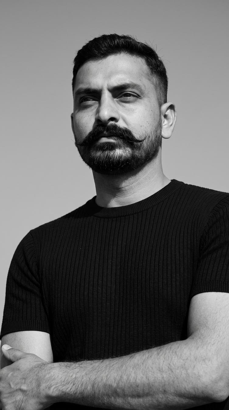 Ultra-realistic black-and-white editorial portrait, low-angle shot, 4:5 aspect ratio. The man fills about 85-90% of the frame in a medium close-up, showing the head, neck, shoulders, upper chest, upper arms, and part of the forearms, with only a small amount of empty space above his head. A handsome athletic man wearing a plain black fitted ribbed crew-neck short-sleeve sweater. His arms are loosely crossed. His body is turned slightly (20°) while his head is turned slightly to the left and tilted upward, with his eyes looking off-camera toward the upper left, giving a calm, confident, thoughtful expression. Strong hard natural sunlight from the upper left illuminates the left side of his hair, forehead, eye, cheek, nose, beard, neck, shoulder, and…