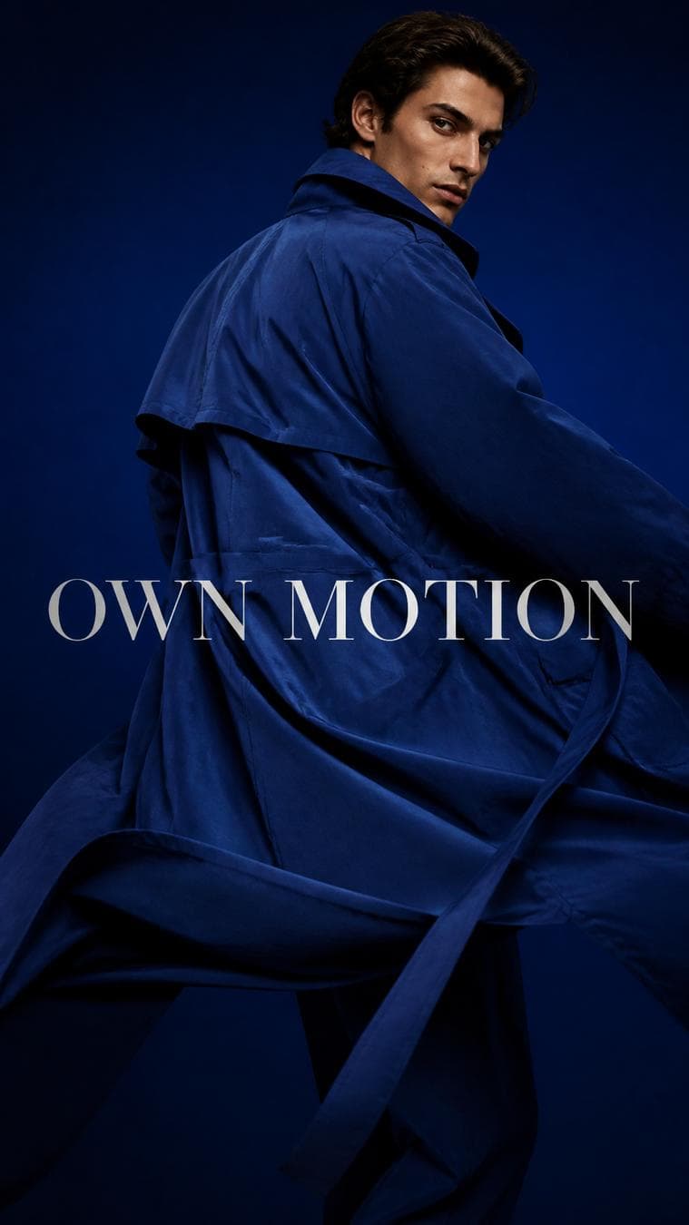 Luxury editorial campaign. Main Subject: Male model wearing an oversized sapphire-blue trench coat. Pose: Half-turn while the coat naturally wraps around the body. Camera: Circular orbit…