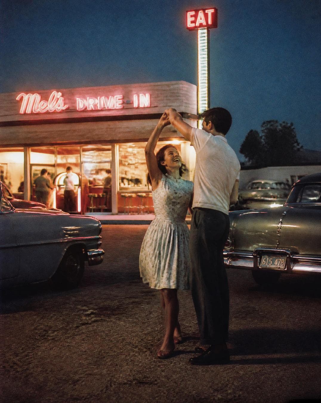1956 roadside diner at blue hour, a young couple spontaneously slow dancing beside a glowing jukebox heard from inside, classic automobiles parked nearby, the woman laughing naturally while the man gently spins her, captured from across the street, candid documentary photography, National Geographic archive, LIFE Magazine 1955, authentic Kodachrome slide film, found family album photograph…
