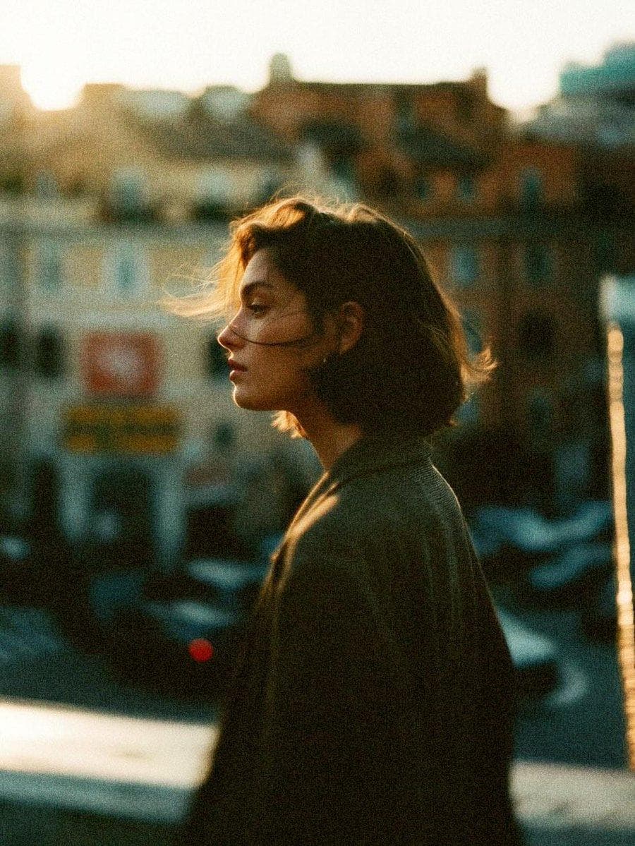 Create a cinematic 35mm analog film photo series featuring the same young adult woman with exact facial identity consistency across every frame. She has short tousled dark-brown bob hair, natural textured skin, subtle freckles, minimal makeup, soft tired eyes, and a quiet, introspective expression. She wears an oversized dark charcoal blazer over a simple dark outfit. Scene: a busy European city street during late golden hour, old urban buildings, parked dark cars, distant pedestrians and traffic, warm sunlight cutting through the streets. The world around her feels fast and restless while she remains emotionally still and detached. IMAGE 1 — SIDE PROFILE: Medium side-profile portrait of the woman standing near the street, facing left. A gentle wind blows loose strands of her short hair across her cheek. Warm sunset rim light outlines her hair and nose. She stares into the distance with a calm, melancholic expression. City architecture becomes heavily blurred behind her. IMAGE 2 — CAR WINDOW CLOSE-UP: Intimate close-up portrait photographed through a slightly dirty car window. Her face appears behind the glass in three-quarter profile, staring outside with distant, thoughtful eyes. Tiny dust particles and subtle marks are visible on the glass. Abstract red and white traffic-light bokeh glows behind her. Reflections softly overlap parts of her face. IMAGE 3 — STREET MOMENT: Candid medium-full street photograph of the same woman beside a parked black car. She turns unexpectedly toward the camera as if someone has just called her name. Wind pushes messy strands across her face. Slight motion blur in the surrounding traffic and pedestrians, spontaneous documentary composition, imperfect framing, emotionally raw. IMAGE 4 — INSIDE CAR: Extreme intimate profile close-up of the same woman sitting inside a car and looking through the side window. Her face is very close to the glass, eyes lowered slightly, lips relaxed. Small droplets, dust and imperfections on the window remain sharply visible while city lights dissolve into red, amber and teal bokeh outside. Quiet loneliness and cinematic stillness. VISUAL STYLE: 1970s–1990s European arthouse cinema, Kodak Portra 800 mixed with Cinestill 800T character, pushed 35mm film, strong organic grain, visible film noise, slightly…