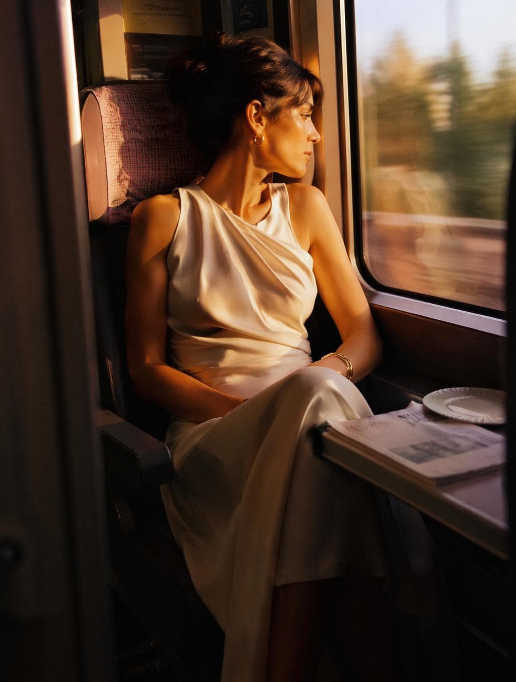 Ultra-realistic cinematic fashion photography of a beautiful woman.Wearing a sophisticated designer dress in soft neutral tones, luxurious fabrics, timeless silhouette.Seated beside a train window during golden hour.Warm sunlight falls across the dress and face.The outside landscape appears blurred by motion.The frame feels accidental, intimate and authentic.Shot through the train cabin as if documenting a fleeting…