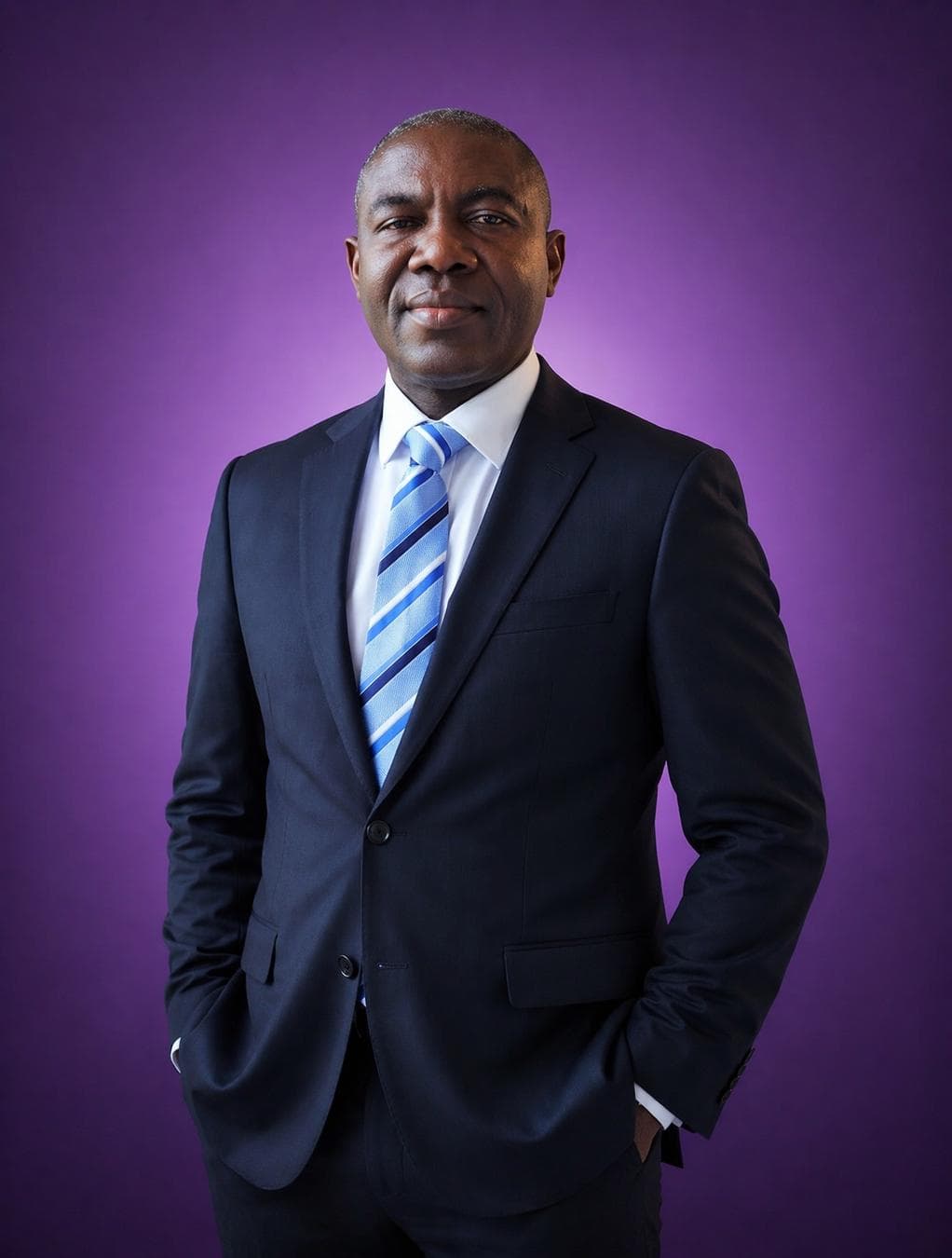 A professional middle-aged man with a dark complexion stands in a centered portrait pose, wearing a tailored charcoal navy wool suit and a crisp white shirt accented by a diagonal blue-striped silk tie. The lighting is a clean, multi-point studio setup that creates soft facial modeling and minimal shadows, emphasizing the fine texture of the fabric and skin. A vibrant, solid violet backdrop provides high-contrast color separation, with a subtle radial gradient emanating from behind the subject to create depth. The composition is a classic corporate headshot, maintaining sharp focus on the eyes and facial features while ensuring the formal attire remains crisp and well-defined against the saturated monochromatic…