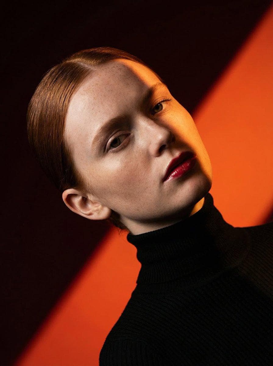 Ultra-high-fashion editorial beauty portrait of a fair-skinned female model with sleek center-parted hair tightly pulled into a low bun, wearing a fitted black ribbed turtleneck sweater. Dramatic studio lighting creates a bold diagonal shadow slicing across the face, emphasizing facial structure, freckles, and glossy deep-red lips. Minimal makeup with natural skin texture, cinematic contrast, rich warm color palette. Background consists of striking geometric color-blocked panels in vibrant orange and deep burgundy red, forming strong diagonal lines. Clean composition, luxury magazine cover aesthetic, contemporary fashion campaign, fine-art portrait photography, sharp focus, ultra-detailed skin, professional studio setup, high dynamic range, moody atmosphere, Vogue-style editorial, medium close-up framing, 85mm…