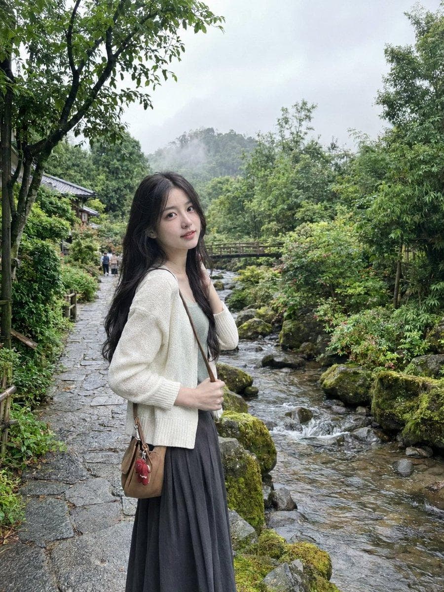 ULTRA-REALISTIC NATURAL SMARTPHONE PHOTOGRAPH, vertical 3:4, spontaneous candid travel portrait of a young East Asian woman standing on a quiet stone walkway beside a misty mountain stream in a lush Japanese-inspired garden. She has long, softly wavy deep-black hair falling naturally over her shoulders and down her back, with a slightly off-center part and a few loose strands gently framing her face. Her facial features are delicate and youthful, with naturally textured skin, subtle realistic pores, minimal makeup, soft pink lips, and a relaxed thoughtful expression as she looks slightly past the camera. She is wearing a cream-colored lightweight knit cardigan over a simple muted sage-green camisole, paired with a flowing dark charcoal pleated midi skirt. A small vintage tan leather crossbody bag rests naturally against her hip, with a tiny red fabric keychain attached to the strap. Her body is angled slightly toward the stream while one foot is naturally positioned a little ahead of the other, as if she has just paused during a quiet morning walk. One hand gently holds the strap of her bag while the other lightly touches a strand of hair near her cheek. The pose feels spontaneous, relaxed, and completely unposed rather than like a fashion shoot. In the background, a clear shallow mountain stream flows over smooth dark stones, surrounded by moss-covered rocks, bamboo, ferns, flowering shrubs, and tall green trees. A small traditional wooden footbridge crosses the stream in the distance, partially hidden by soft vegetation. A couple of tiny distant pedestrians are naturally present on the pathway, adding subtle scale without distracting from the subject. The atmosphere is cool and slightly misty after a light morning rain. The sky is mostly covered with pale gray clouds, creating soft diffused natural daylight with gentle shadows and realistic muted green tones. Small droplets of…