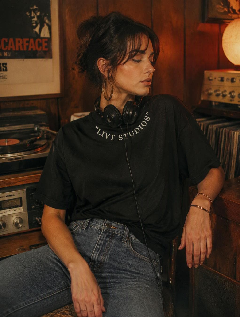 Use the uploaded images as visual references.REFERENCE IMAGE 1: use it as the exact reference for the WOMAN, her pose, facial features, hairstyle, body proportions, clothing fit, hands, accessories, camera angle and overall composition.REFERENCE IMAGE 2: use it as the exact reference for the LIVT CLUB T-SHIRT.Replace ONLY the woman’s existing T-shirt with the exact black LIVT CLUB T-shirt shown in reference image 2.Do not change anything else in the original composition.T-SHIRT — ABSOLUTE ACCURACYAnalyze the second reference image carefully before generating.Reproduce the exact garment:black heavyweight cotton T-shirtoversized relaxed fitsame collarsame sleeve length and widthsame shoulder proportionssame fabric weight and texturesame black colorsame natural wrinkles and foldssame garment constructionThe shirt must look physically worn by the woman, not pasted onto her.The white text around the collar must remain exactly:“LIVT STUDIOS”including the quotation marks and the curved placement around the neckline.Do not invent, modify, misspell, remove or add any letters.The collar print must follow the natural curvature of the neckline and the fabric.The shirt must naturally conform to the woman’s:shoulderschestwaistarmsposebody movementPreserve realistic fabric tension, shadows, folds and contact with the body.The garment must look like a real premium LIVT CLUB product photographed in the scene.LIVT CLUB VISUAL IDENTITYMake the finished image feel fully integrated into the LIVT CLUB universe.Preserve the warm 1970s-inspired editorial atmosphere:dark wooden wallsvintage record playervinyl recordsold stereo equipmentretro posterswarm tungsten lightingburnt orange, brown, cream and deep black palettesubtle analog film grainsoft halationnatural photographic imperfectionscinematic editorial lightingThe image should feel like an authentic 1970s-inspired LIVT CLUB fashion editorial, not a modern photo with a vintage filter.WOMANKeep the woman from the original reference image exactly as she is.Preserve:facehairstylebangsfacial expressionskin tonebody proportionsposearm positionhandsjewelryjeansheadphonescamera…