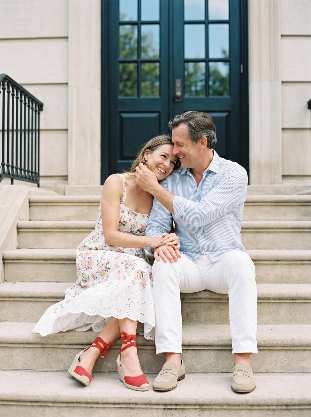 On a quiet afternoon, a relaxed, romantic moment unfolds on the steps of a classical building: the man in a light striped linen shirt, white trousers and beige loafers; the woman, in her late 40s, in a white floral sundress with eyelet hem and red ankle-tie espadrille wedges. Keep the background realistic—broad limestone steps, iron railing, tall dark door with mullioned glass reflecting faint greenery and sky. Action: start from the seated pose; the woman rests her head on his shoulder, they interlace fingers, share a small laugh, he gently brushes a strand of hair from her face, she adjusts her ankle tie with a slight foot movement, a light breeze lifts the dress hem, they glance to camera then back to each other, ending with a warm forehead-to-forehead touch. Style: cinematic, photorealistic, professional film look; natural skin tones, realistic fabric motion, subtle film grain, creamy background bokeh. Camera: medium two-shot at eye level on 35 mm with a slow 5–8% dolly-in; intercut 85 mm close-ups on hands and smiles; a brief 50 mm tilt from their shoes up to faces; one rack focus from the doorway to the couple; maintain 24 fps, ~1/48 shutter. Composition: centered symmetry with rule-of-thirds facial placement; leading lines from steps; clean headroom; include at…