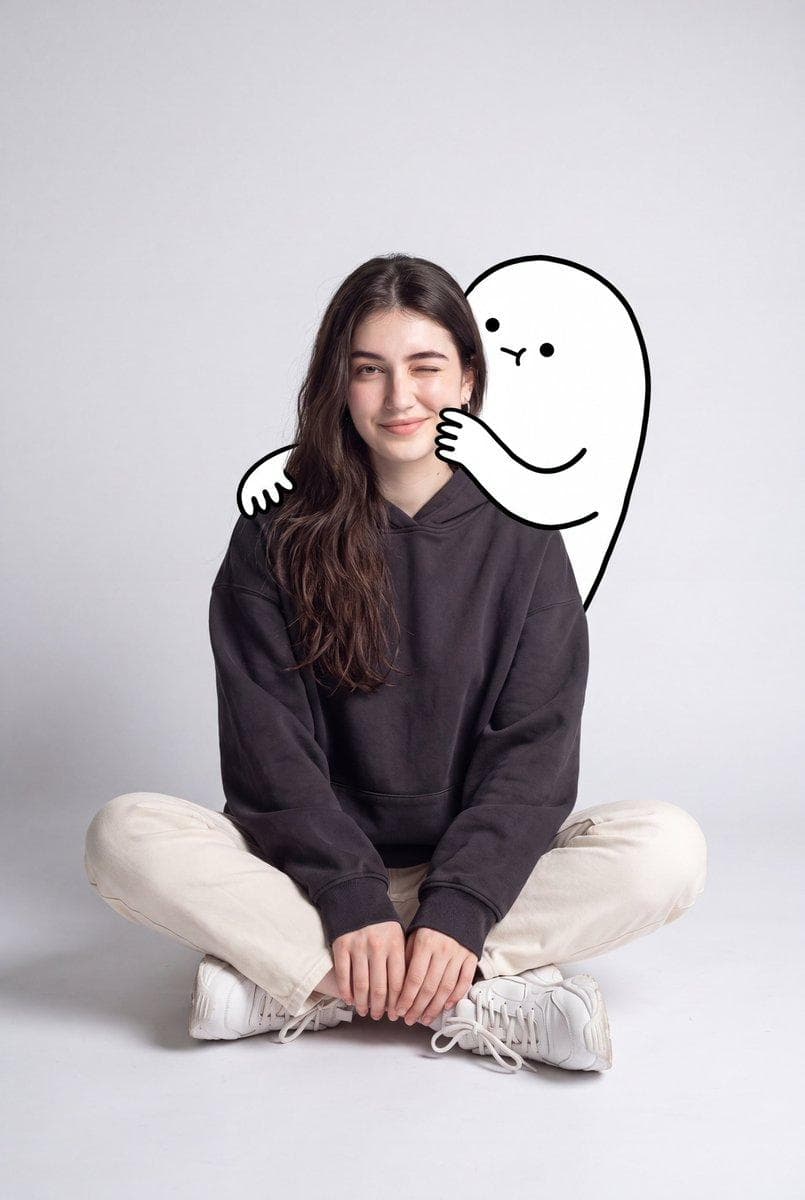 A cute minimalist portrait of a young woman sitting casually on the floor in a clean studio setting with a light grey background. She wears an oversized dark charcoal hoodie, cream straight-leg pants, white socks, and chunky white sneakers. Relaxed seated pose with crossed legs, one eye slightly closed in a playful wink and a soft smile. Behind her, a large black-and-white doodle character hugs her from behind, wrapping its cartoon arms around her shoulders and gently squeezing her cheek.The doodle figure has a simple round head, tiny eyes, and a…