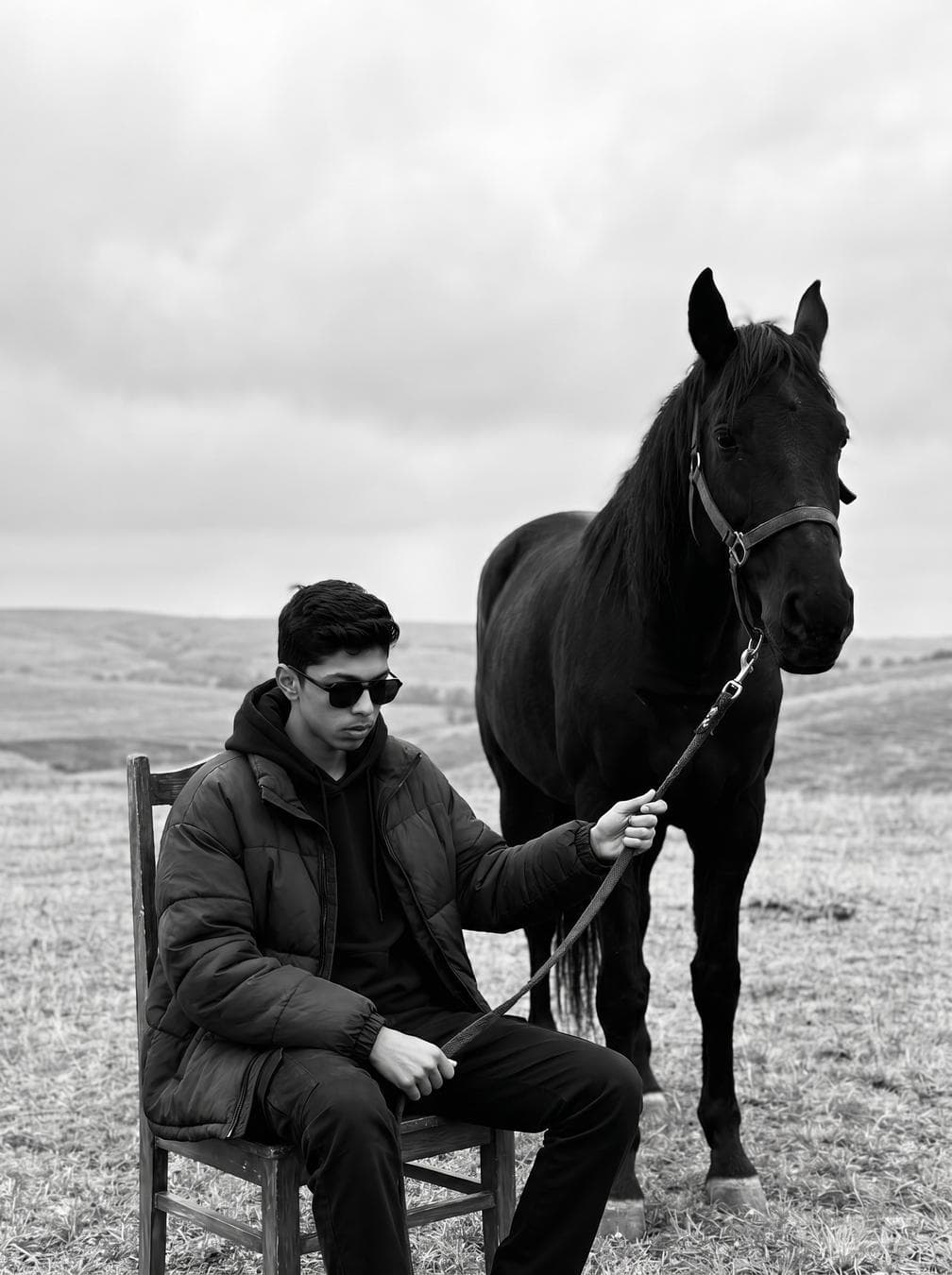 { "model": "Nano Banana Pro", "scene": "Open field landscape under an overcast sky, minimal natural surroundings", "subject": { "type": "Young man", "pose": "Seated on a wooden chair, legs relaxed apart, holding a horse lead calmly", "expression": "Detached, introspective, controlled", "gaze": "Looking downward, partially obscured by sunglasses", "emotion": "Quiet dominance, solitude, inner gravity" }, "appearance": { "hair": { "color": "Dark", "style": "Short, slightly tousled curls" }, "facial_features": { "beard": "Light stubble", "accessories": ["Dark sunglasses"] }, "skin": "Natural texture, matte finish" }, "attire": { "outerwear": "Oversized dark padded jacket", "innerwear": "Dark hoodie", "pants": "Relaxed-fit dark trousers", "footwear": "Black leather boots", "style_note": "Minimalist, masculine, utilitarian fashion" }, "secondary_subject": { "type": "Black horse", "position": "Standing beside the man, head lowered slightly", "interaction": "Held calmly by a loose lead", "symbolism": "Strength, instinct, controlled power" }, "environment": { "location": "Open grassy field", "background": [ "Rolling hills", "Soft distant landscape", "No urban elements" ], "atmosphere": "Quiet, isolated, contemplative" }, "lighting": { "type": "Natural diffused daylight", "conditions": "Overcast sky", "effect": "Soft shadows, low contrast, even tonal range" }, "camera": { "angle": "Eye-level to slightly low-angle", "lens": "50mm cinematic portrait lens", "depth_of_field": "Moderate, subject and horse in focus, background gently blurred", "framing": "Vertical, full-body portrait with symbolic spacing" }…
