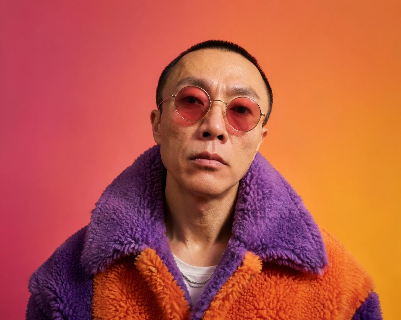 A stylish skinhead man from the reference, poses in a high-fashion aesthetic, wearing circular rose-tinted sunglasses and an oversized, textured sherpa coat featuring a bold violet and orange color-blocked design. The subject is positioned against a vibrant, seamless gradient backdrop shifting from warm magenta to golden yellow, creating a clean studio isolation effect. Soft, diffused lighting wraps around the subject’s facial features, emphasizing subsurface scattering on the skin while minimizing harsh shadows. The thick, tactile pile of the synthetic fleece absorbs light, providing a rich contrast to the translucent, reflective surface of the rose-colored lenses. The composition is a sharp, centered portrait that prioritizes saturated color harmony…