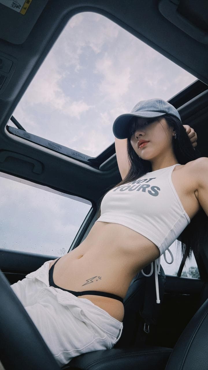 Vertical low-angle candid photo inside car, cool tone film texture, cute cool East Asian girl, long straight black hair, faded grey baseball cap covering half eyes, tight white cropped halter tank top with grey letter print, slim flat toned abdomen with small tattoo on waist, white low-waist casual pants, thin black underwear strap exposed, perfect S waist and hip curve, shot under car sunroof, cloudy pale sky in background, dark car frame framing shot, soft…