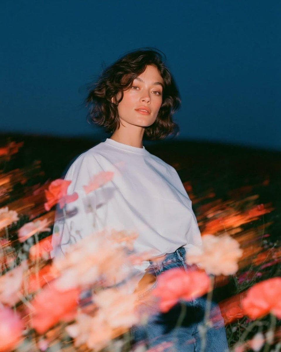 Create a cinematic fashion editorial portrait at blue hour in an open wildflower field. A beautiful young woman with short, softly wavy dark hair and natural makeup stands among blooming pink and white poppies, wearing an oversized white crew-neck T-shirt tucked into high-waisted vintage blue jeans. She gazes directly into the camera with a calm, effortless expression while a gentle breeze moves her hair. Photograph using a slow shutter with rear-curtain flash, keeping the subject perfectly sharp while the flowers in the foreground create glowing motion-blur streaks and painterly light trails sweeping diagonally across the frame. Warm flash illumination freezes the model against a rich deep navy twilight sky, creating a striking contrast between cool ambient light and warm floral highlights. Foreground filled with heavily blurred flowers, soft bokeh, translucent ghosting, glowing streaks, dreamy long-exposure movement, and subtle lens bloom. Background fades into a dark meadow with minimal distractions, emphasizing the…
