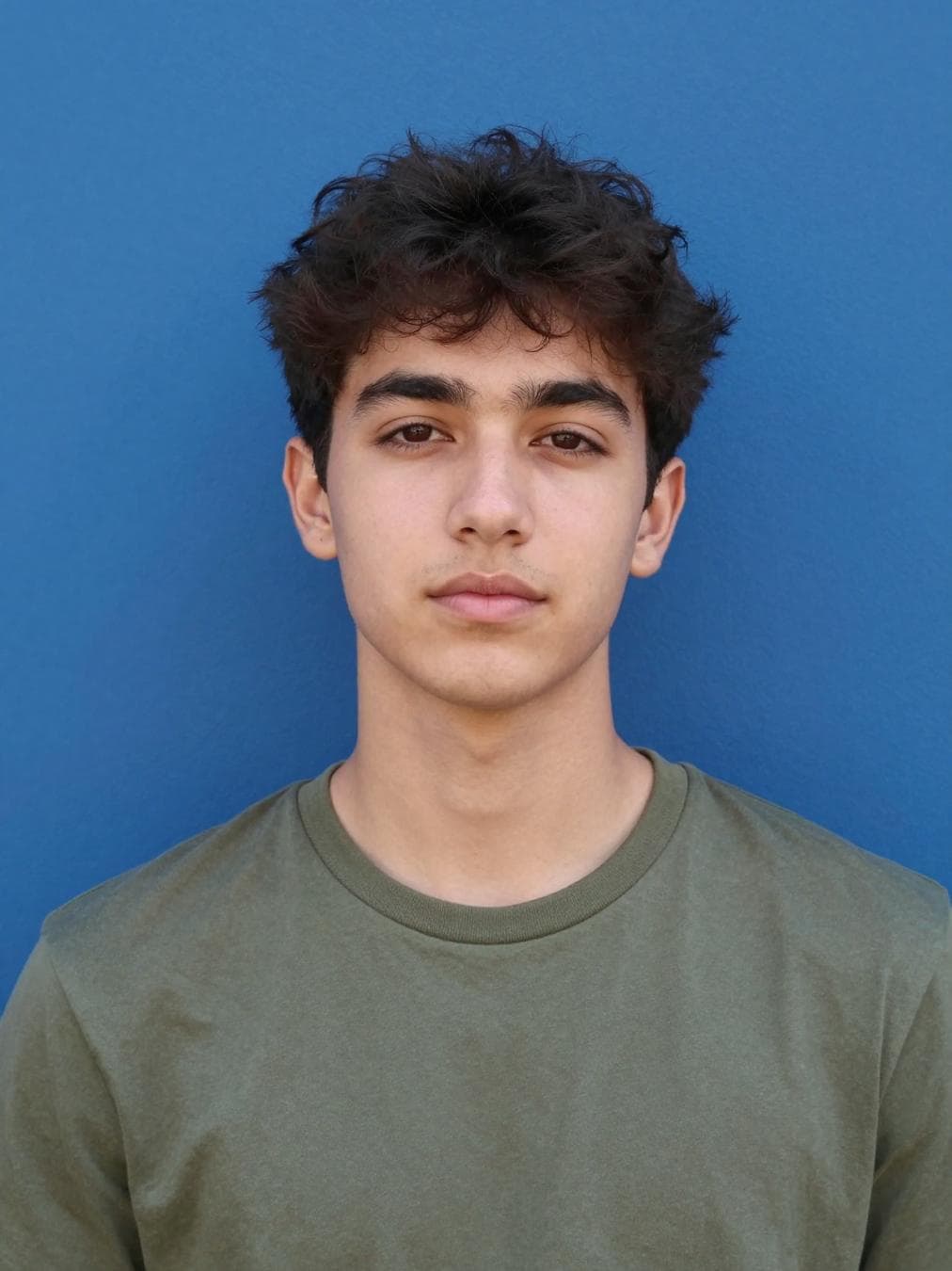 A young man with dark, textured hair poses for a centered portrait against a flat, vibrant ultramarine blue wall. The lighting is soft and diffused, likely from an overhead interior source, resulting in subtle shadows beneath the eyes and chin while maintaining natural skin tones with minimal specular highlights. The subject wears a simple olive-green crew-neck t-shirt, providing a muted color contrast against the saturated background. The composition is a direct, eye-level head-and-shoulders shot, characterized by a slight atmospheric haze or soft-focus effect typical of front-facing mobile device photography. The background remains uniform and out of focus, emphasizing the subject's neutral expression and the clean, high-contrast separation between the…