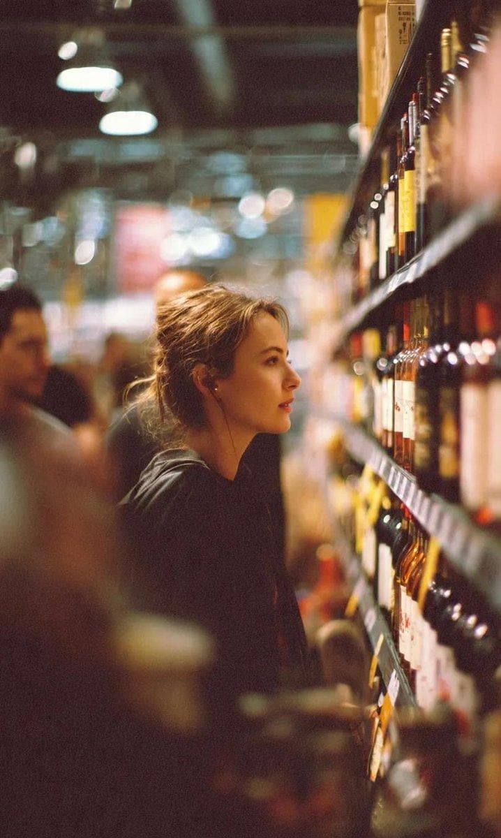 A cinematic side-profile portrait of a young woman standing in a grocery store aisle, carefully looking at a shelf filled with wine bottles. She has her hair tied in a loose messy bun and wears a simple dark outfit. The environment is softly lit with warm indoor lighting, creating a cozy, realistic atmosphere. The shelves are filled with neatly arranged bottles, while other shoppers appear blurred in the background. Shallow depth of field with…