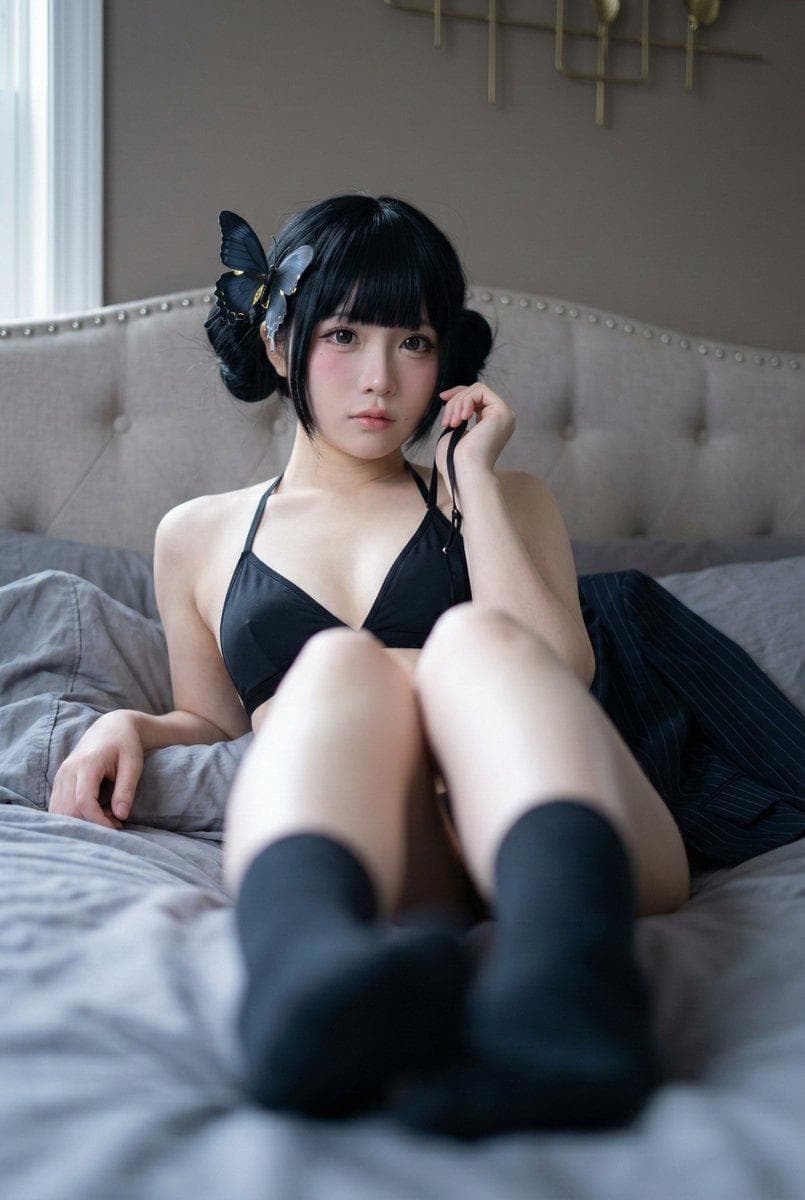 { "subject": { "description": "An adult woman in a cosplay-inspired boudoir portrait, reclining on a bed with foreshortened legs toward the camera, soft doll-like makeup, and a distinctive blue-black butterfly hair clip.", "age": "adult, early-to-mid 20s", "expression": { "eyes": { "look": "wide, glossy contact-lens look", "energy": "quiet and slightly guarded", "direction": "looking directly at the camera" }, "mouth": { "position": "gently closed, slightly pressed", "energy": "subtle, calm" }, "overall": "soft, intimate, just-awake stillness" }, "face": { "preserve_original": true, "makeup": "cosplay doll makeup: smooth base, soft pink blush, subtle contour, defined eyeliner, natural pink lips" }, "hair": { "color": "jet black", "style": "straight blunt bangs with twin bun silhouette and long side sections framing the face", "effect": "sleek, slightly glossy strands with a few loose flyaways" }, "body": { "frame": "slim, petite", "waist": "narrow", "chest": "moderate", "legs": "long with strong perspective toward the lens", "skin": { "visible_areas": "shoulders, arms, upper chest, abdomen, thighs, feet", "tone": "very fair with cool undertone", "texture": "soft-touch, lightly retouched skin with gentle natural gradients", "lighting_effect": "smooth highlight roll-off, faint shadow shaping under collarbones and along the torso" } }, "pose": { "position": "reclining on the bed, torso angled slightly to the right, legs extended toward the camera", "base": "one hand holding a thin black strap loosely, the other hand with an index finger resting near the cheek", "overall": "intimate, relaxed, slightly teasing but non-explicit" }, "clothing": { "top": { "type": "black triangle halter bikini/lingerie top with thin straps", "color": "black", "details": "minimal coverage, clean edges, thin string straps", "effect": "sharp strap lines contrasting with soft bedding folds" }, "bottom": { "type": "matching black bikini/lingerie bottom (mostly obscured by pose and framing)", "color": "black", "details": "not clearly visible; implied coordinated set" } } }, "accessories": { "headwear": "large butterfly hair clip on the left side (blue-black wings with warm gold accents)", "prop": "a dark pinstripe blazer draped near the body, thin black strap held in hand" }, "photography": { "camera_style": "realistic cosplay boudoir portrait, high-resolution digital look with mild skin retouching", "angle": "low angle from the foot of the bed, slight tilt, strong foreshortening", "shot_type": "three-quarter body portrait with foreground foot blur and face in sharp focus", "aspect_ratio": "2:3 vertical", "texture": "clean detail, soft micro-contrast, subtle grain, no heavy sharpening", "lighting": "soft diffused window light from camera-left, gentle shadow falloff, cool gray mood", "depth_of_field": "shallow depth of field, foreground (feet/legs) noticeably blurred, eyes and face crisp" }, "background": { "setting": "quiet bedroom scene on a bed", "wall_color": "warm taupe-gray", "elements": [ "tufted upholstered headboard with diamond pattern", "rumpled gray duvet/blanket and pillows", "partial metallic wall decor near the top edge" ], "atmosphere": "hushed, private, late-afternoon calm", "lighting"…