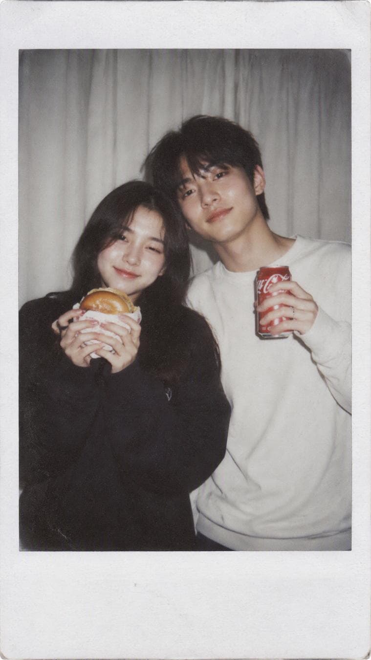 Subject and interaction Create a realistic Polaroid-style candid photograph of the two young Asians couple shown in the reference images. Preserve both people’s facial identity, features, expressions, and proportions exactly; do not alter, beautify, or distort their faces. They stand close together in a relaxed, playful pose, leaning slightly toward each other. The girl holds a burger, and the boy holds a red Coca-Cola can. Their expressions and body language feel natural, friendly, and unposed. Composition Eye-level medium shot with both people centred in the frame. Keep the framing casual and slightly imperfect, like an ordinary snapshot taken spontaneously indoors. Plain white curtains fill the background. Lighting and atmosphere A single direct on-camera flash illuminates the pair against a dim indoor room, creating the characteristic bright frontal flash and gentle light scatter of an instant-camera photo. Keep the mood cosy, intimate, and nostalgic, with realistic exposure falloff toward the edges. Visual style and texture Authentic classic Polaroid instant-film appearance: subtle film grain, soft focus, slightly softened edges…