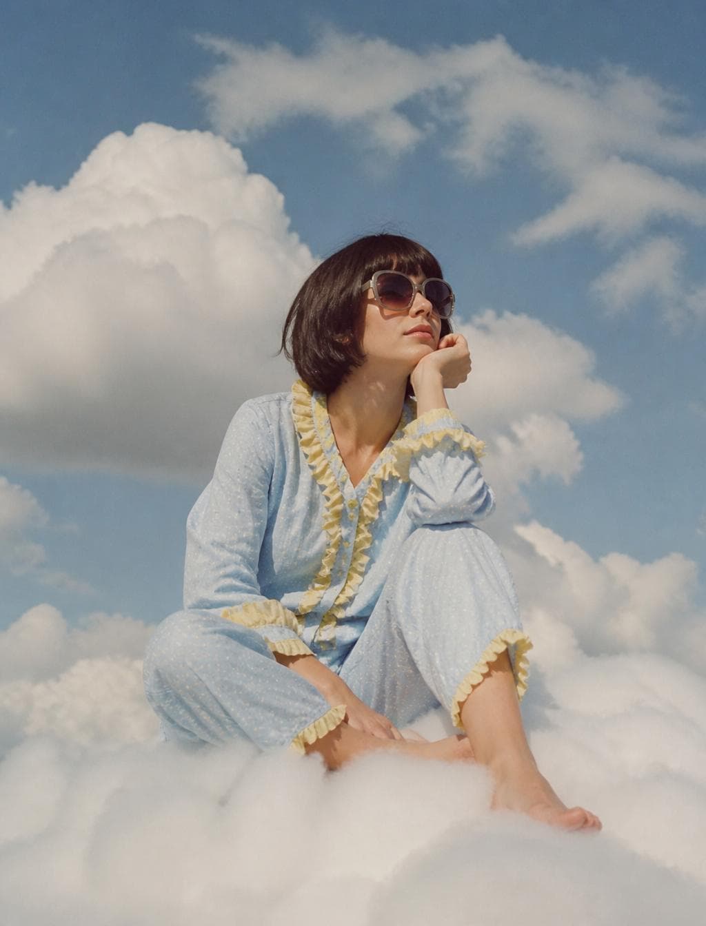 Photo of an eccentric woman sitting on top of white fluffy clouds, wearing large sunglasses and light blue pajamas with yellow ruffles, she has short dark hair in a bob cut…