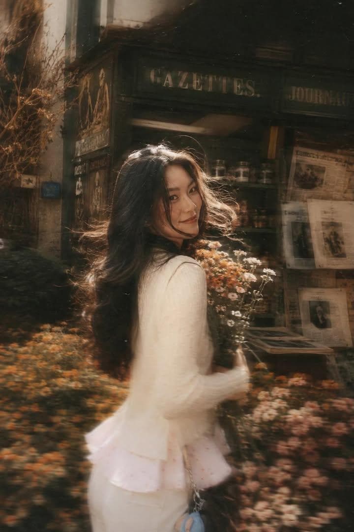 A beautiful young woman with long dark flowing hair holding a bouquet of soft pink roses, turning back and looking at the camera while walking through a flower-filled European street, vintage newspaper kiosk in background, warm golden hour sunlight, dreamy atmosphere, soft glow, cinematic backlighting, wind blowing through hair, shallow depth of field, creamy bokeh, pastel color palette, romantic Parisian aesthetic, delicate white…