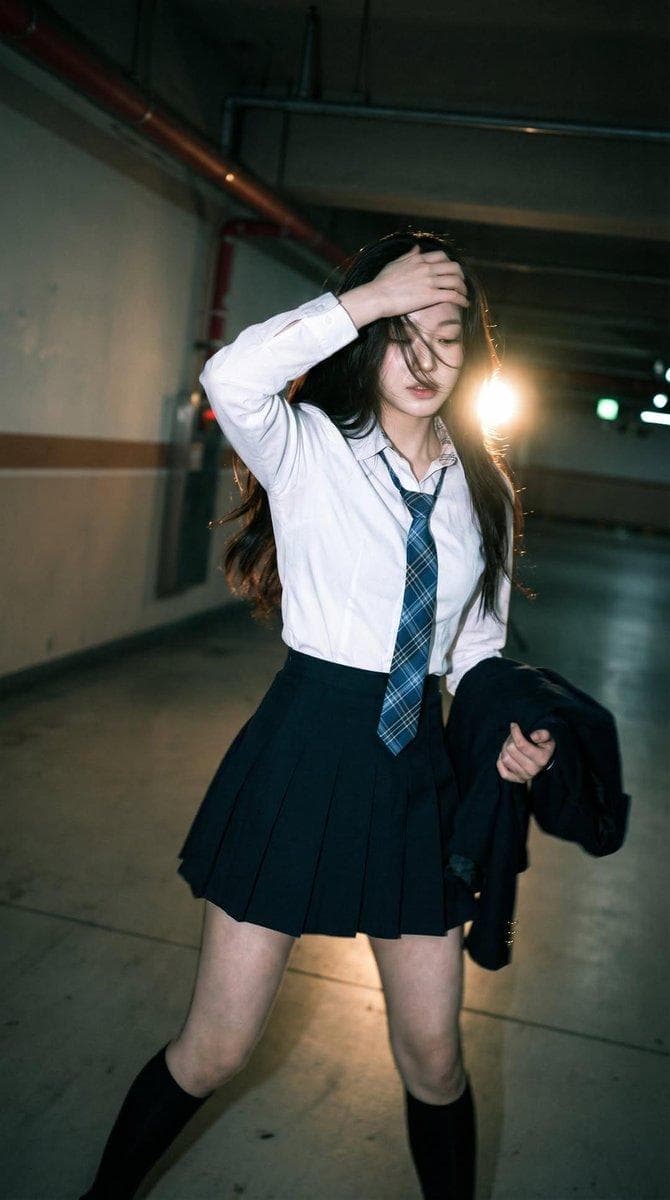 Candid portrait of a young woman in a dim parking garage, wearing a white shirt with a blue plaid tie and pleated mini skirt, black knee-high socks, outfit moving naturally with motion, captured mid-step with one knee slightly bent and torso subtly twisted, creating a dynamic silhouette, one arm raised to partially cover her face while long hair falls across it, the other hand holding a jacket at…